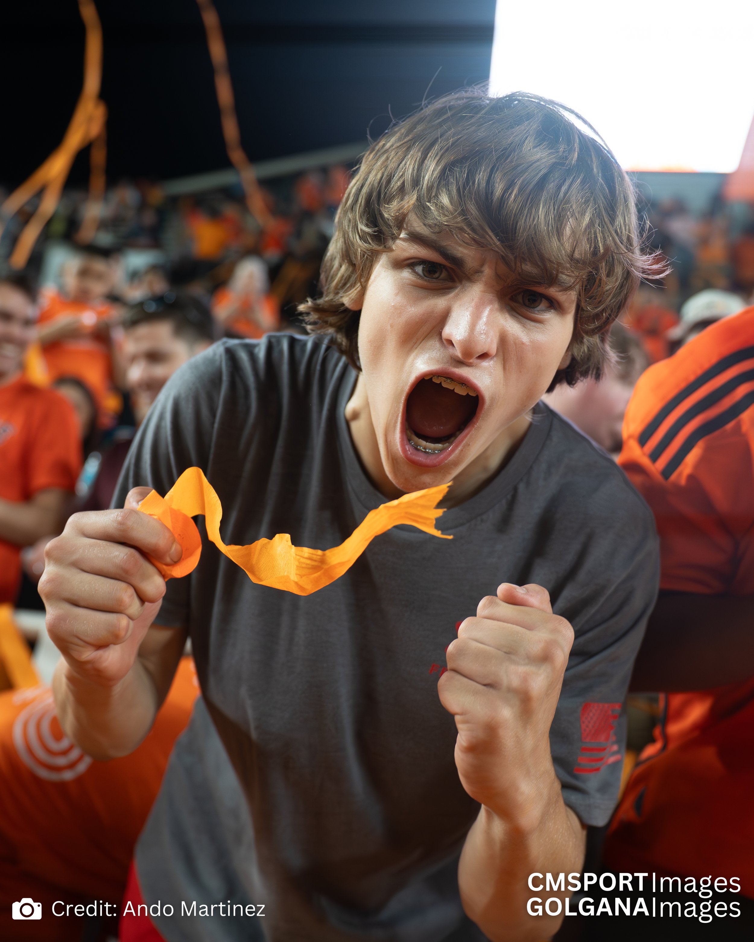 A young man at a sports event, cheering with his fists clenched and mouth open, wearing a gray sports shirt, with a crowd in the background dressed in orange.