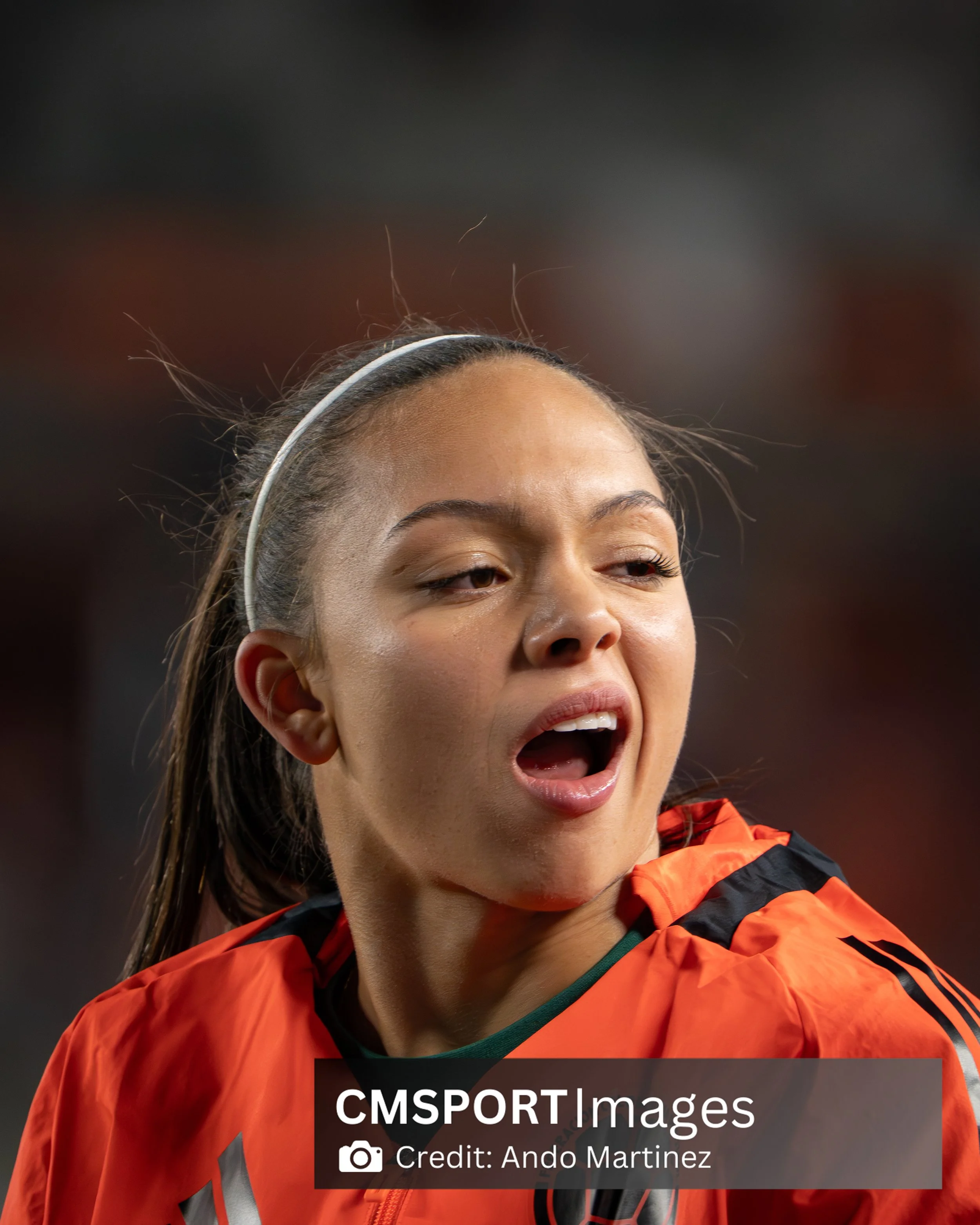Close-up of a female soccer player with an open mouth, wearing an orange sports jersey and a white headband, during a game.