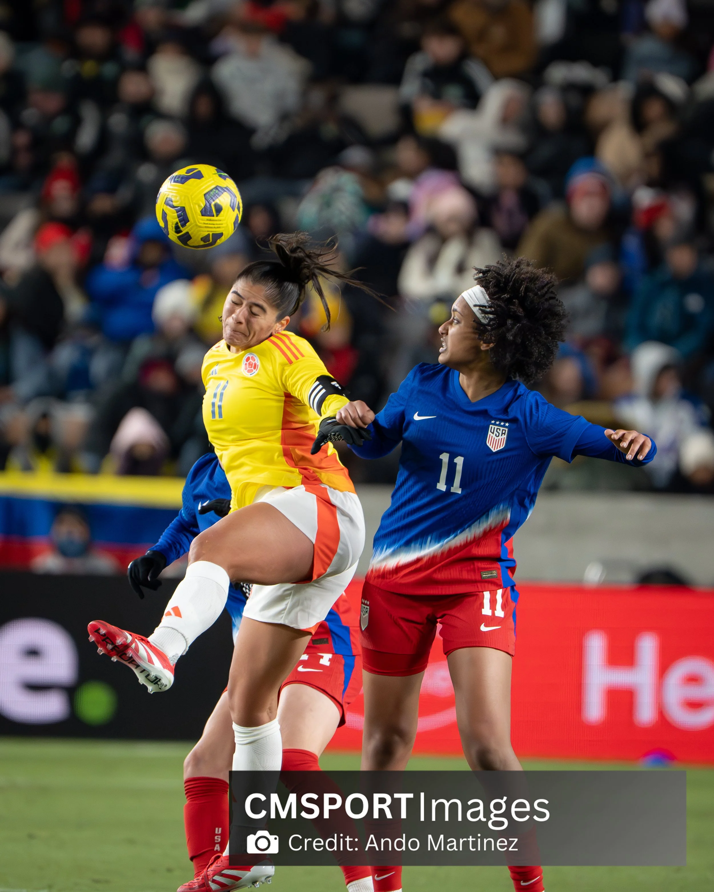 Two female soccer players, one in a yellow jersey and one in a blue jersey, compete for the ball during a match with a crowd in the background.
