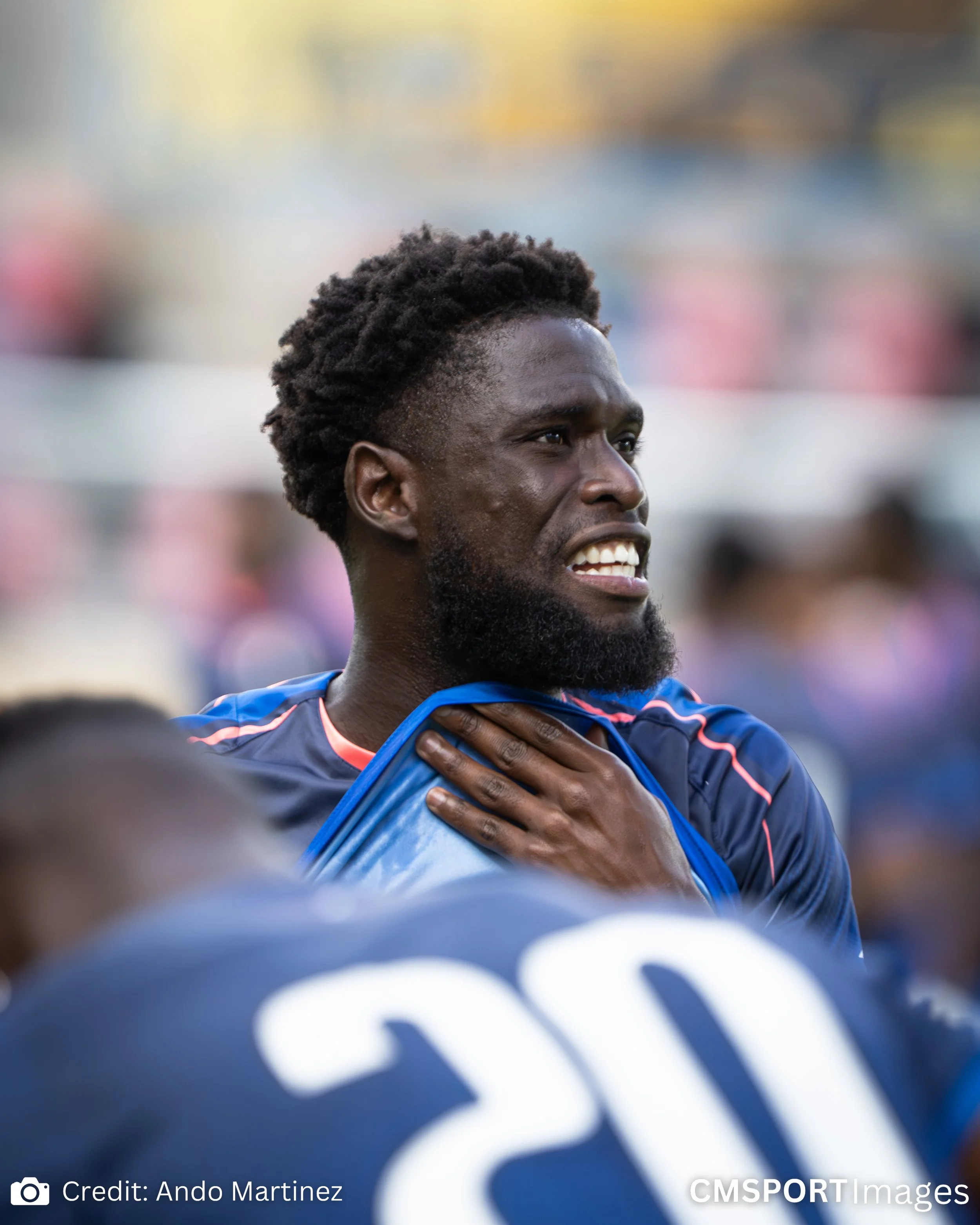 A male athlete with a beard and styled hair, wearing a sports jersey, holding his neck with one hand, on a sports field during a game.