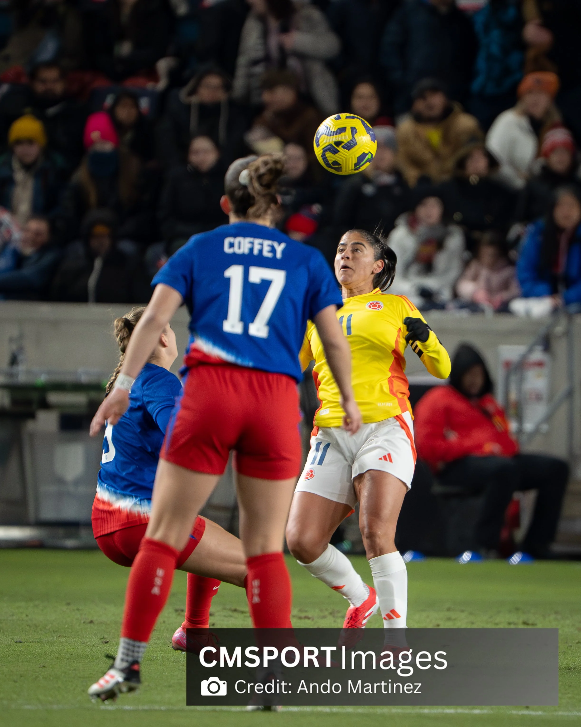 Two female soccer players, one in a yellow jersey and the other in a blue jersey with the number 17, compete for the ball during a match with a crowd watching in the background.
