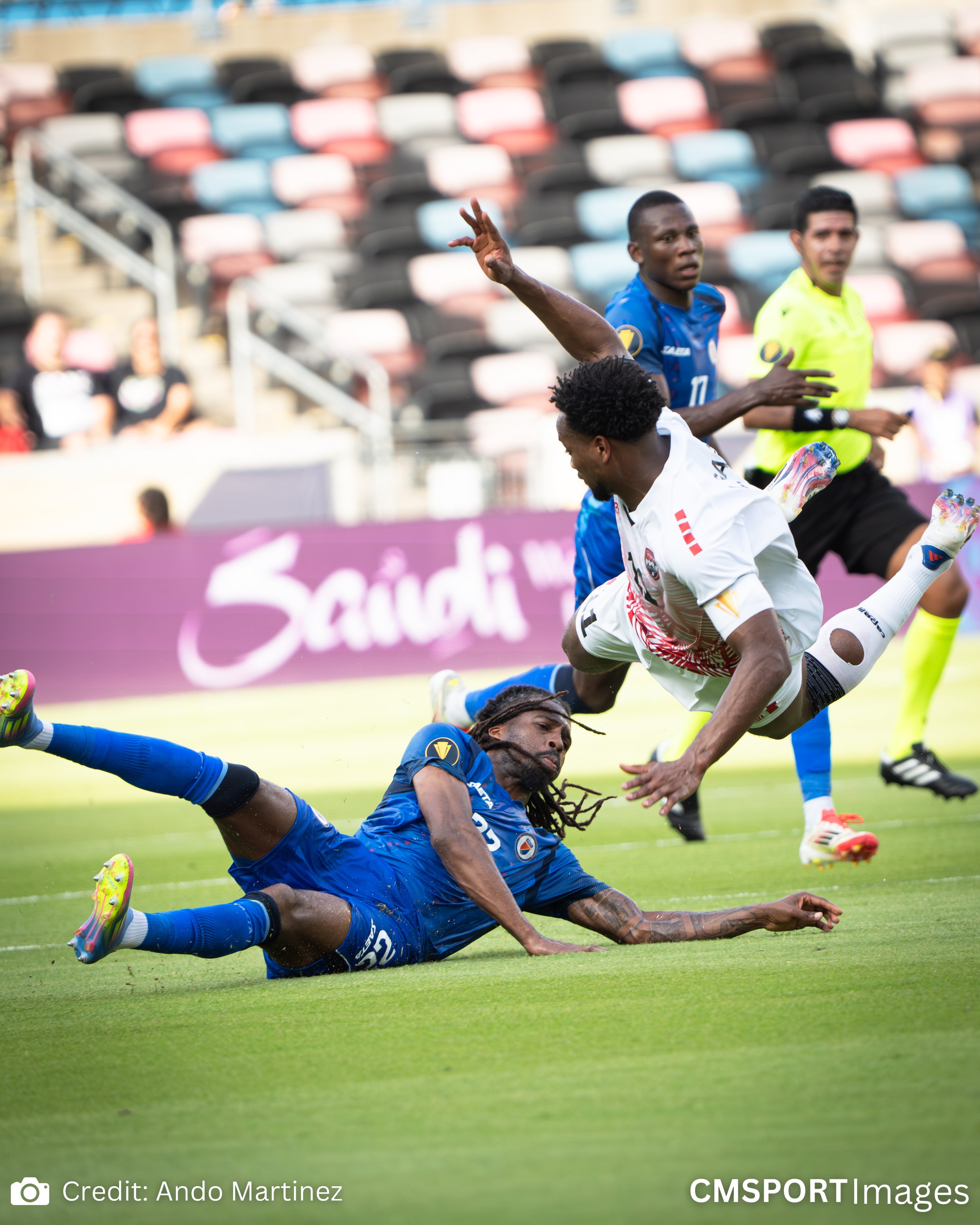 A soccer match with one player in blue falling on the ground and another player in white nearly upside down trying to avoid contact. A referee is observing the action in the background.