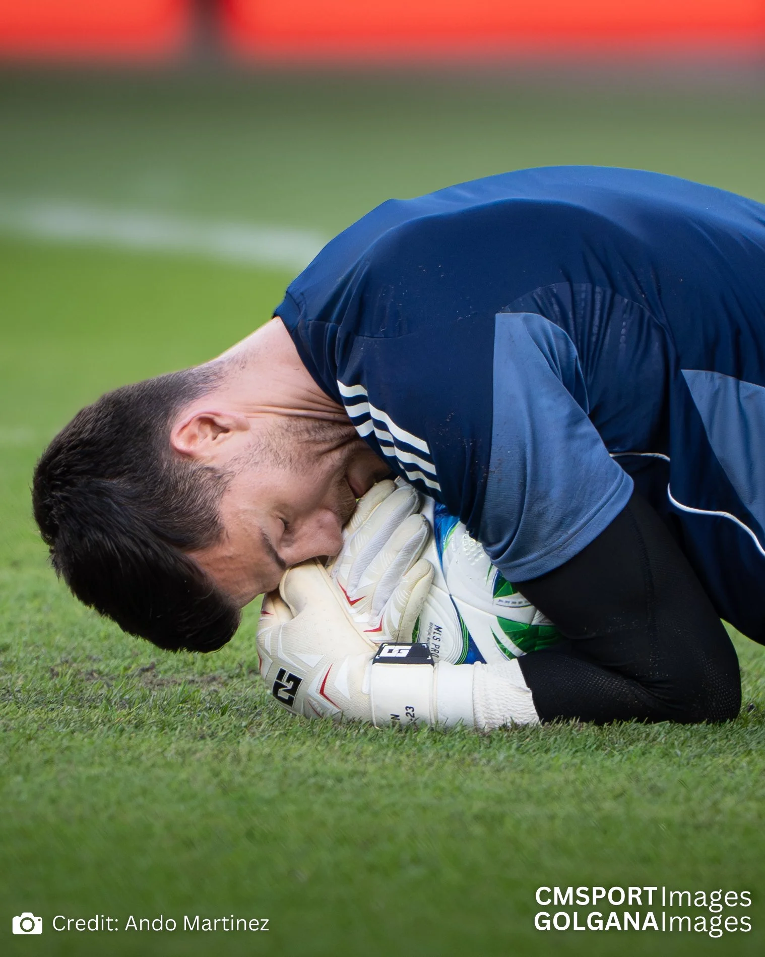 A soccer goalkeeper lying on the grass with his eyes closed, clutching a soccer ball near his face, wearing a blue jersey and white gloves, during a game.