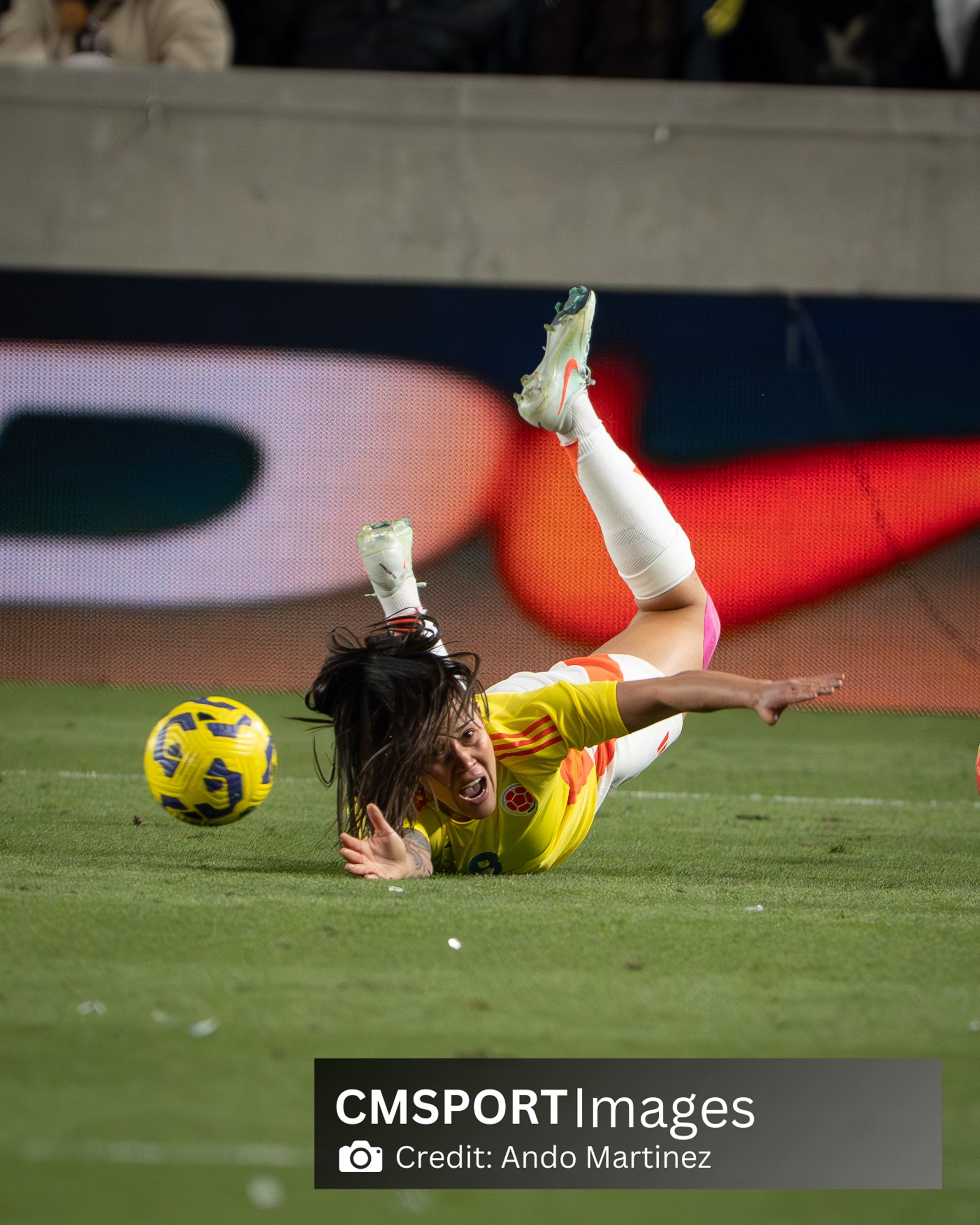 A female soccer player in a yellow jersey is falling to the ground after missing a ball, with her body stretched out, her face showing an intense expression, and her right arm extended on the grass. She wears white socks and cleats, and her long hair