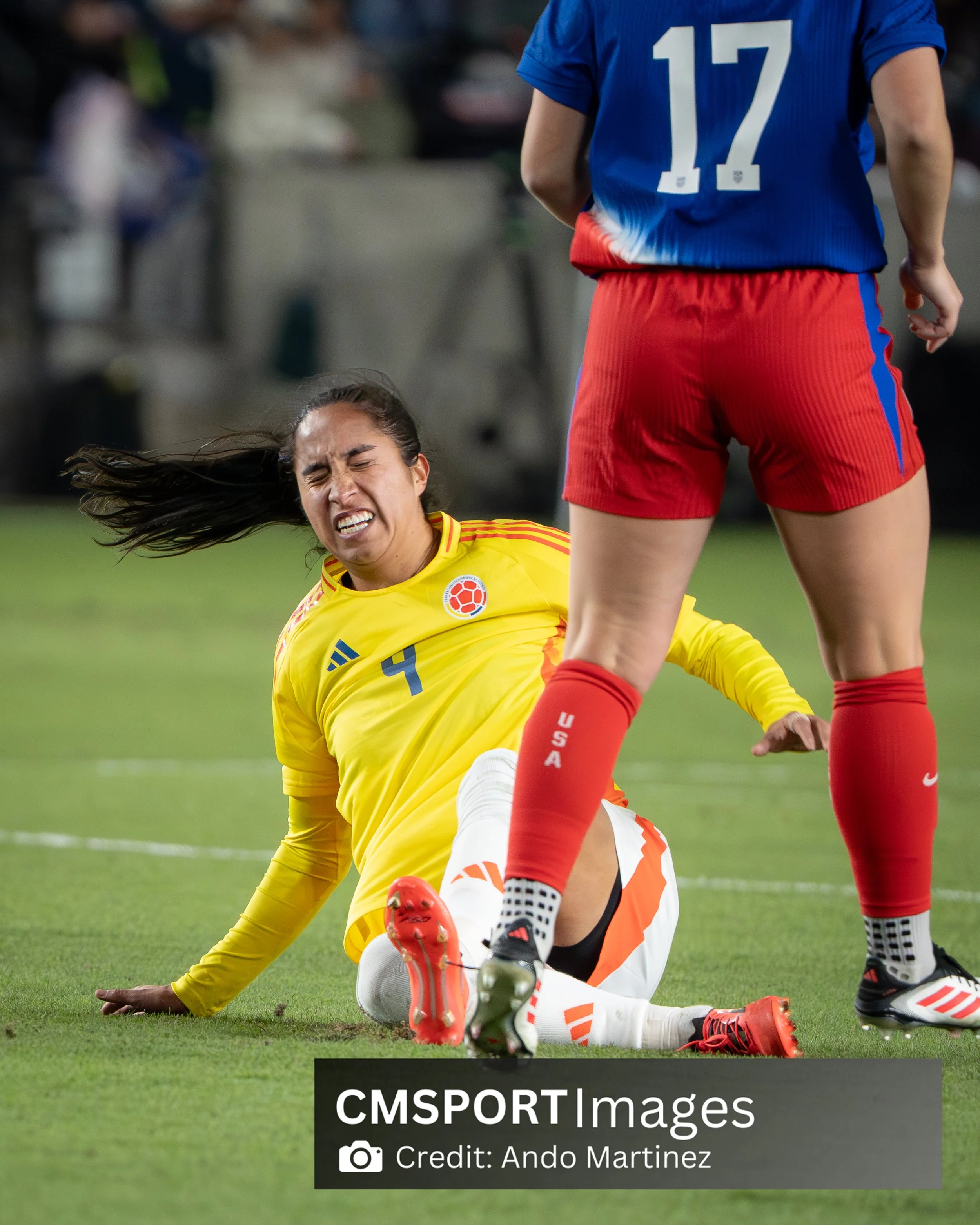 A female soccer player in a yellow Colombia jersey is on the ground, grimacing in pain, while another female player in a red USA uniform stands nearby on the field.