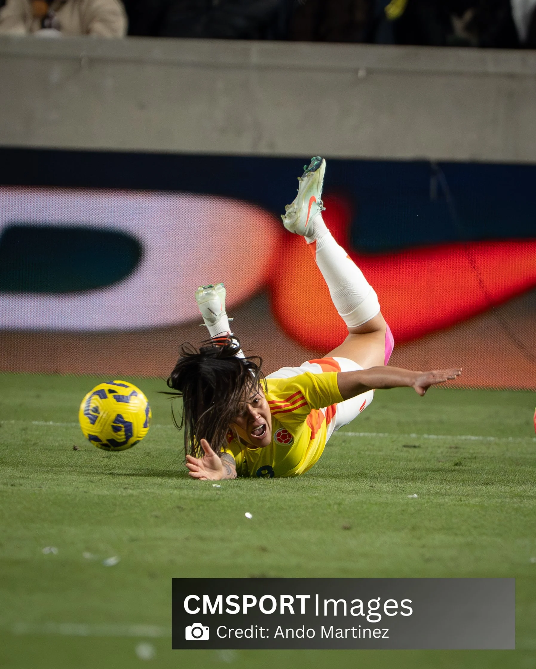 A female soccer player in a yellow uniform is falling or diving onto the grass field after an attempt to reach a yellow and blue soccer ball. Her hair is loose, and her facial expression shows effort or frustration. The background features a digital 