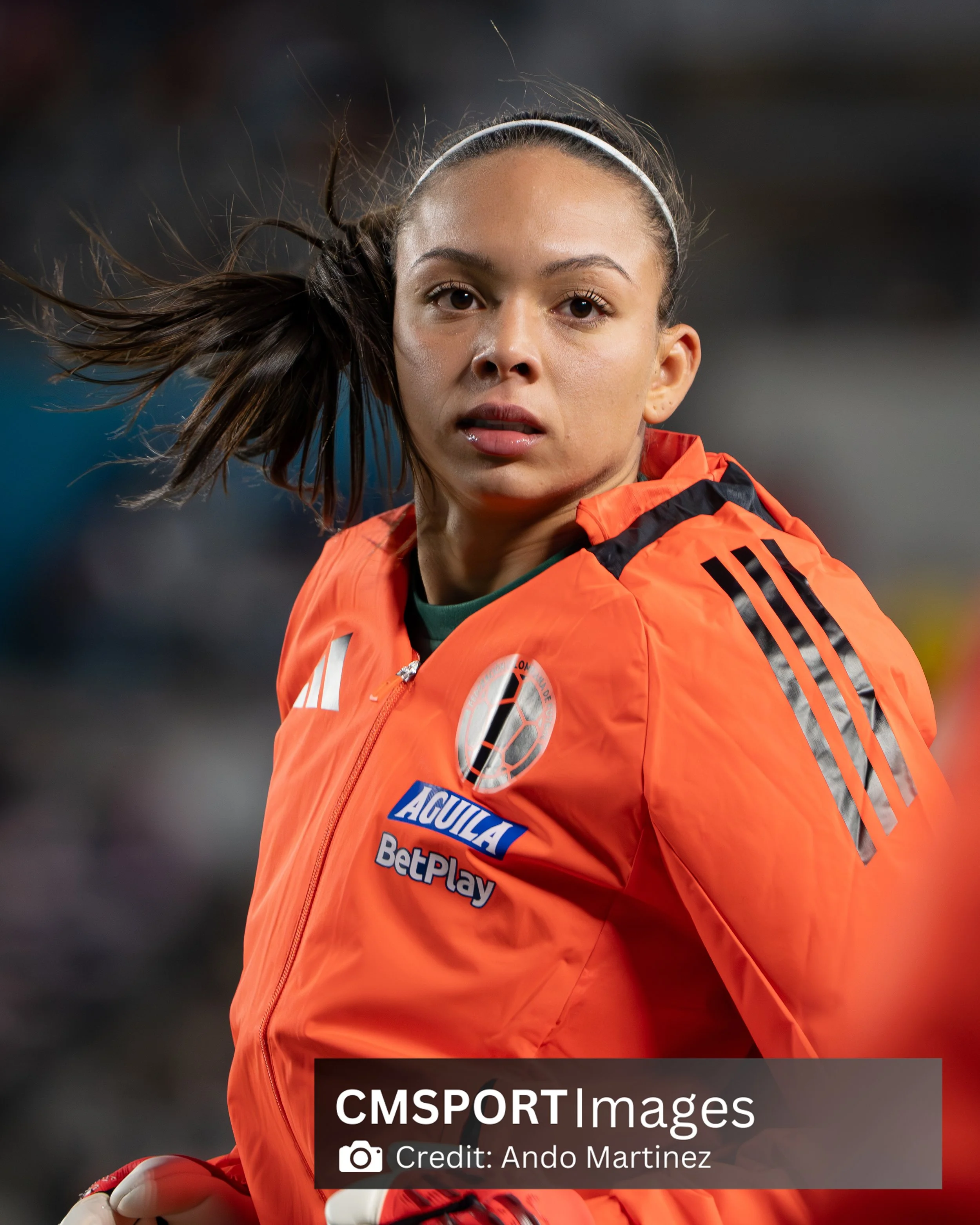Female soccer player with brown hair tied back, wearing an orange jacket with sponsor logos, on the field during a game.