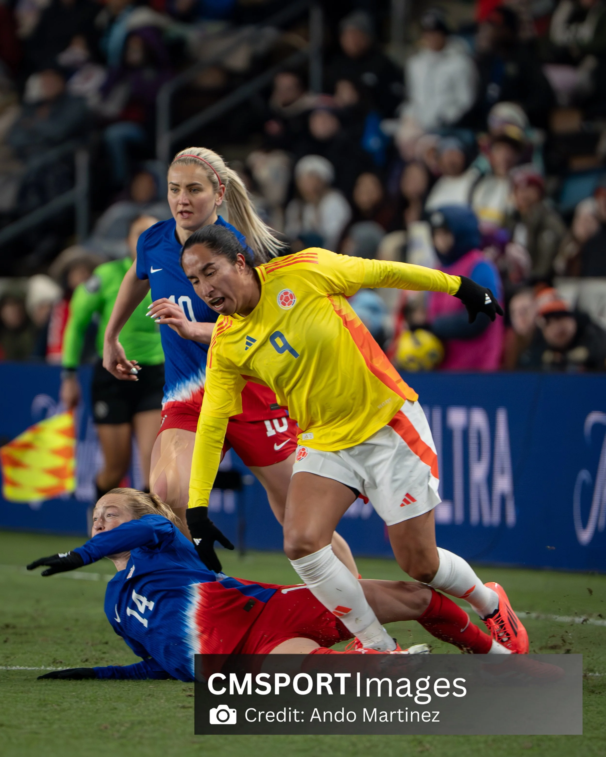 Two female soccer players, one in a blue and red uniform and another in a yellow and white uniform, are competing for the ball during a match. The player in yellow is falling while the player in blue is on the ground, and other players and spectators