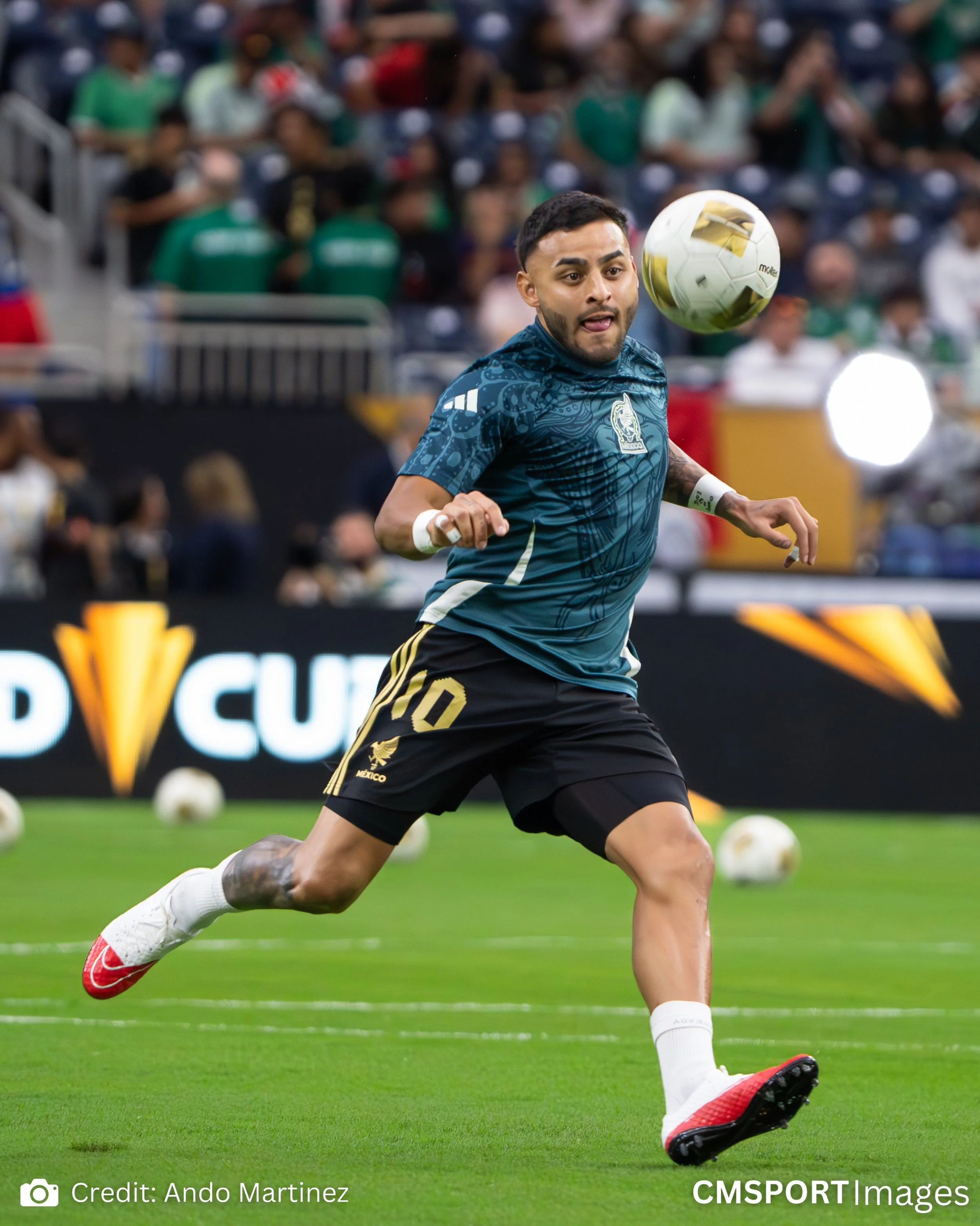A soccer player wearing a dark blue jersey with the number 10 on his shorts, black shorts, and white shoes with red soles, is running on a green field during a match, focused on a soccer ball in the air.