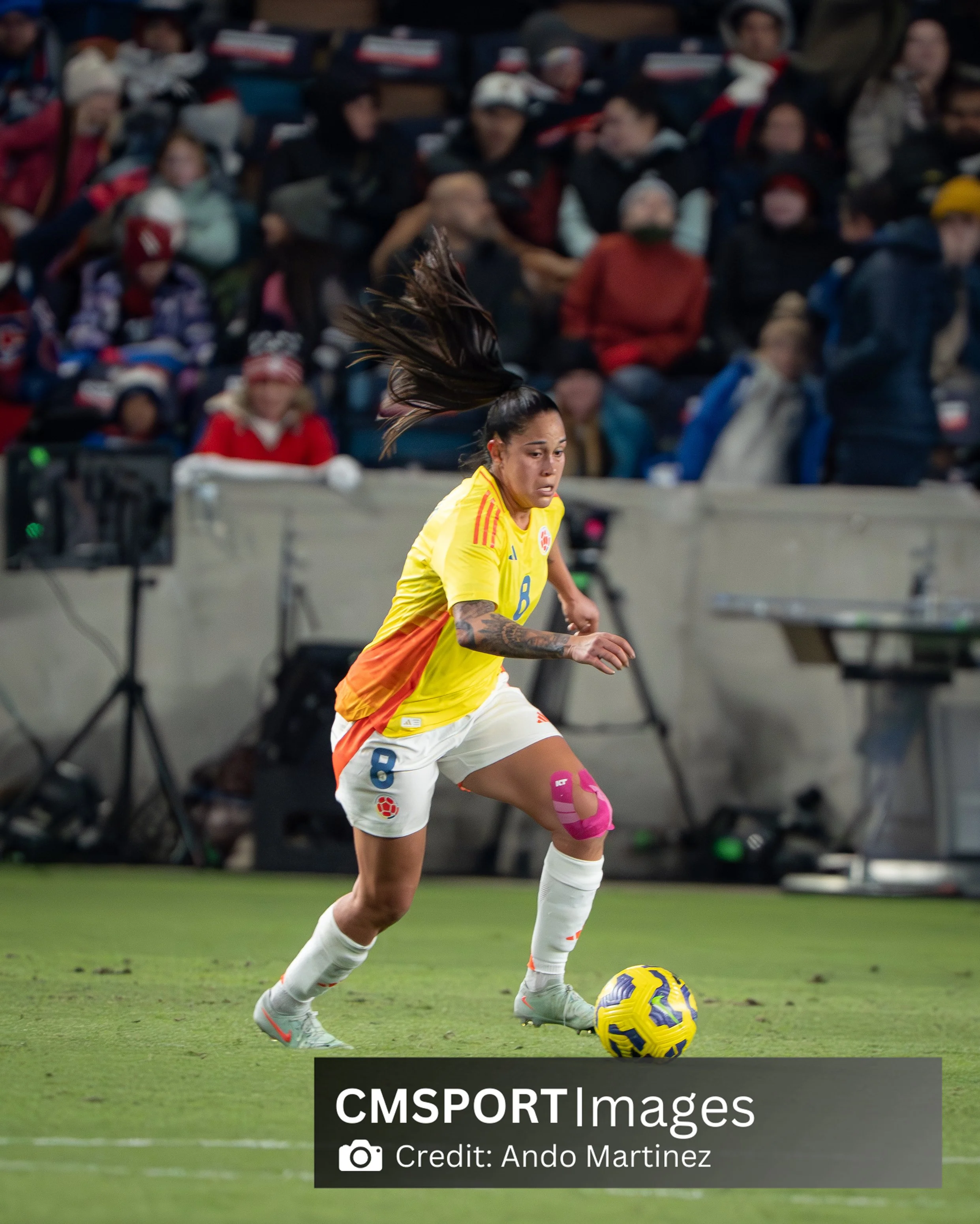 A female soccer player in a yellow jersey with the number 8 on it, dribbling a yellow and purple soccer ball on the field, with a crowd of spectators in winter clothing in the background.