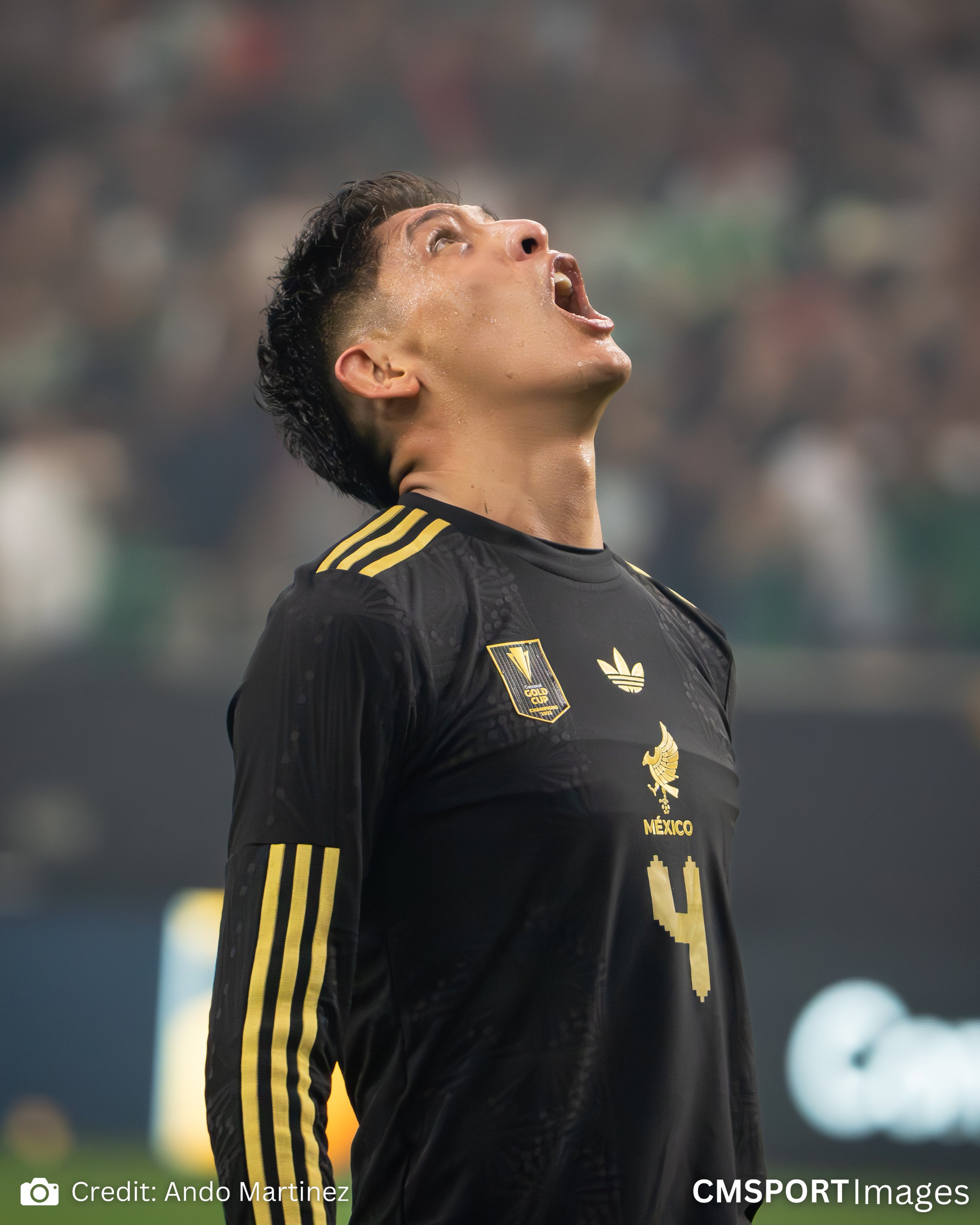 Soccer player wearing a black Mexico national team jersey with gold accents, looking upwards with mouth open, celebrating during a match.
