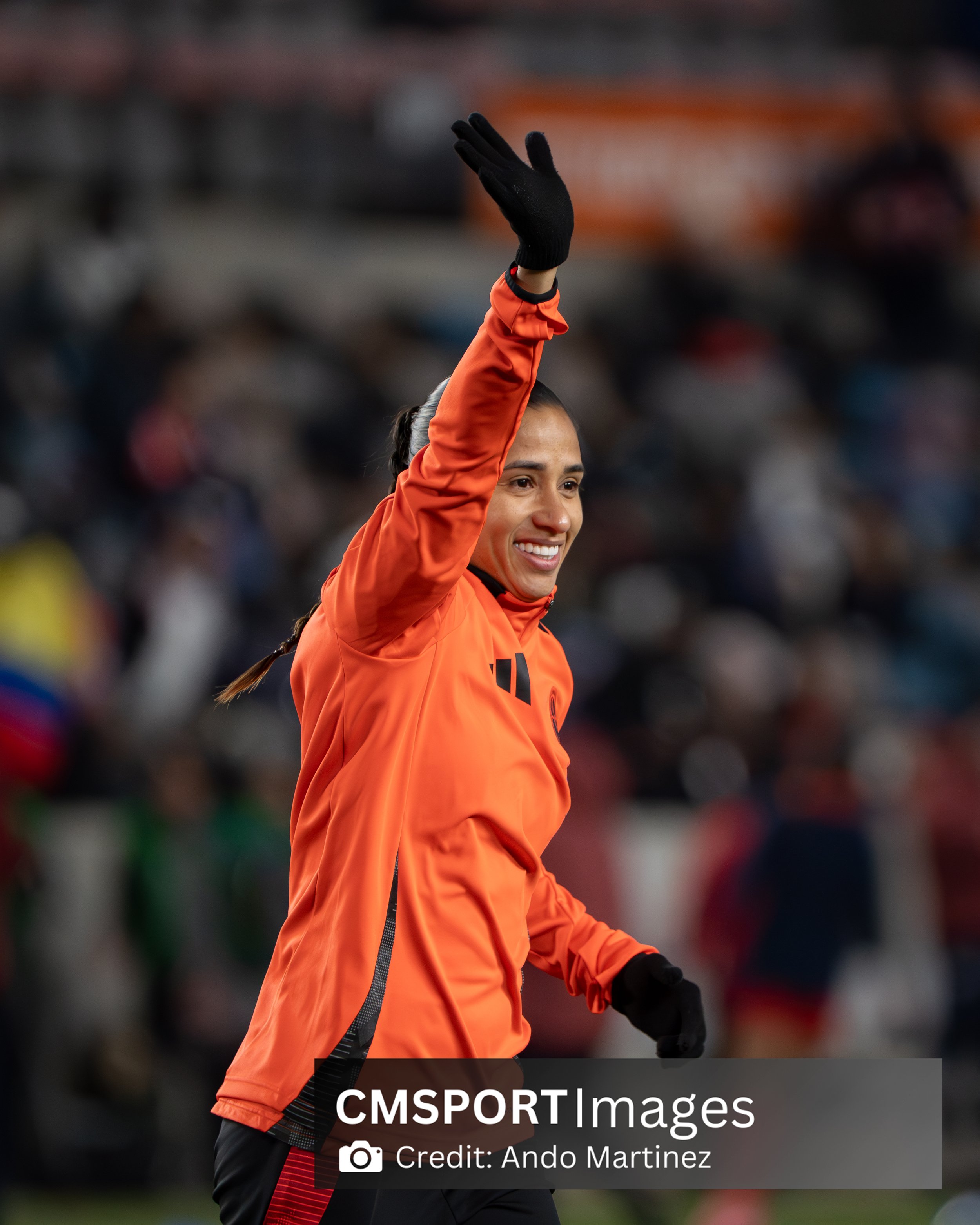 A female soccer player wearing an orange jersey and black gloves waving to the crowd at a stadium.