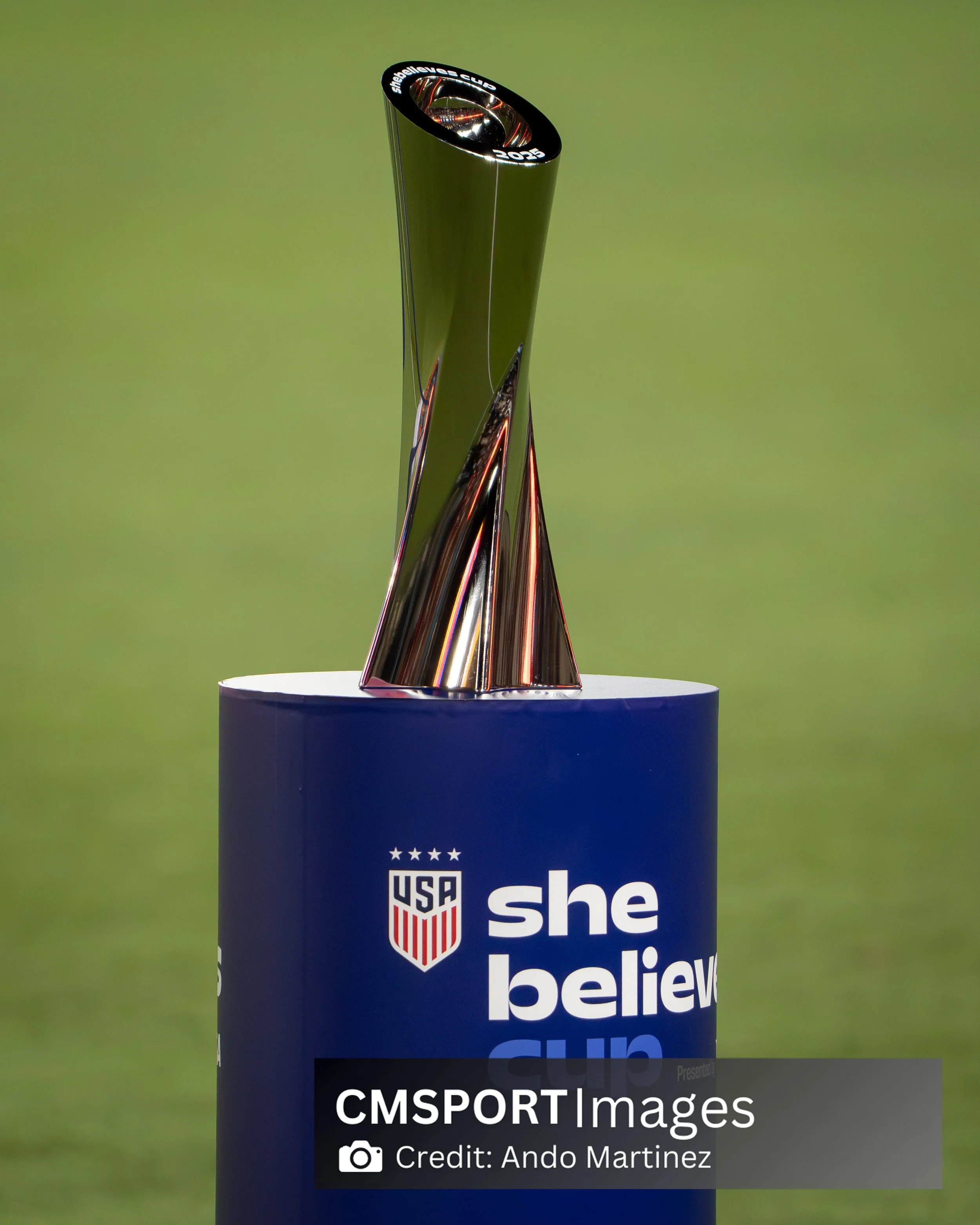 A trophy with a metallic finish atop a blue stand with the US Soccer logo and the phrase "she believes" printed on it.