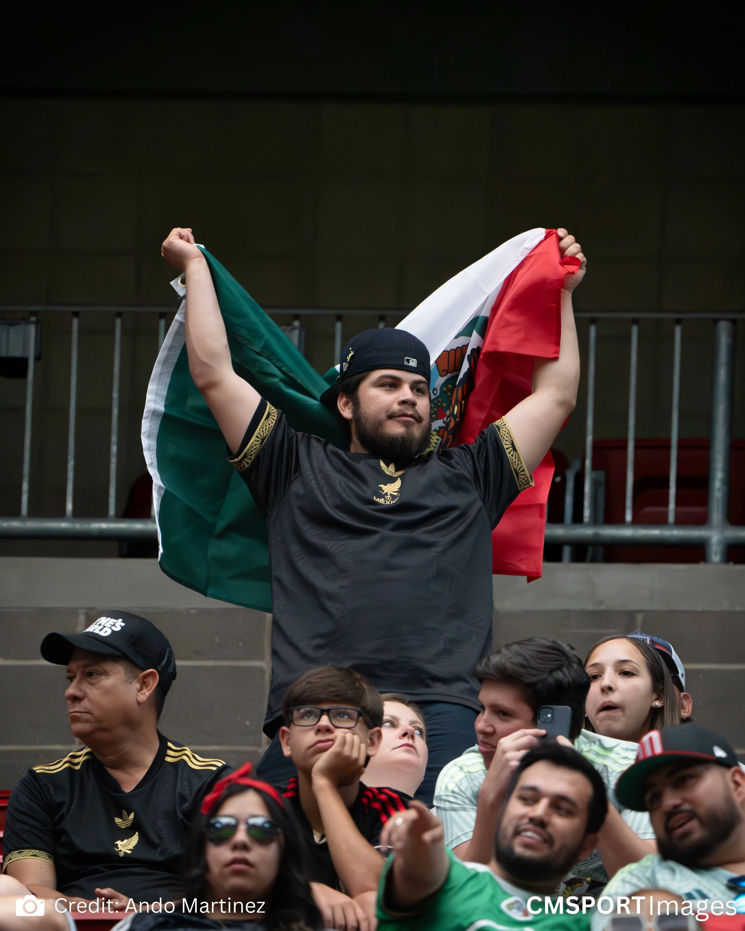 A man stands on a stadium seat holding a Mexican flag behind his head, with others watching in the foreground.