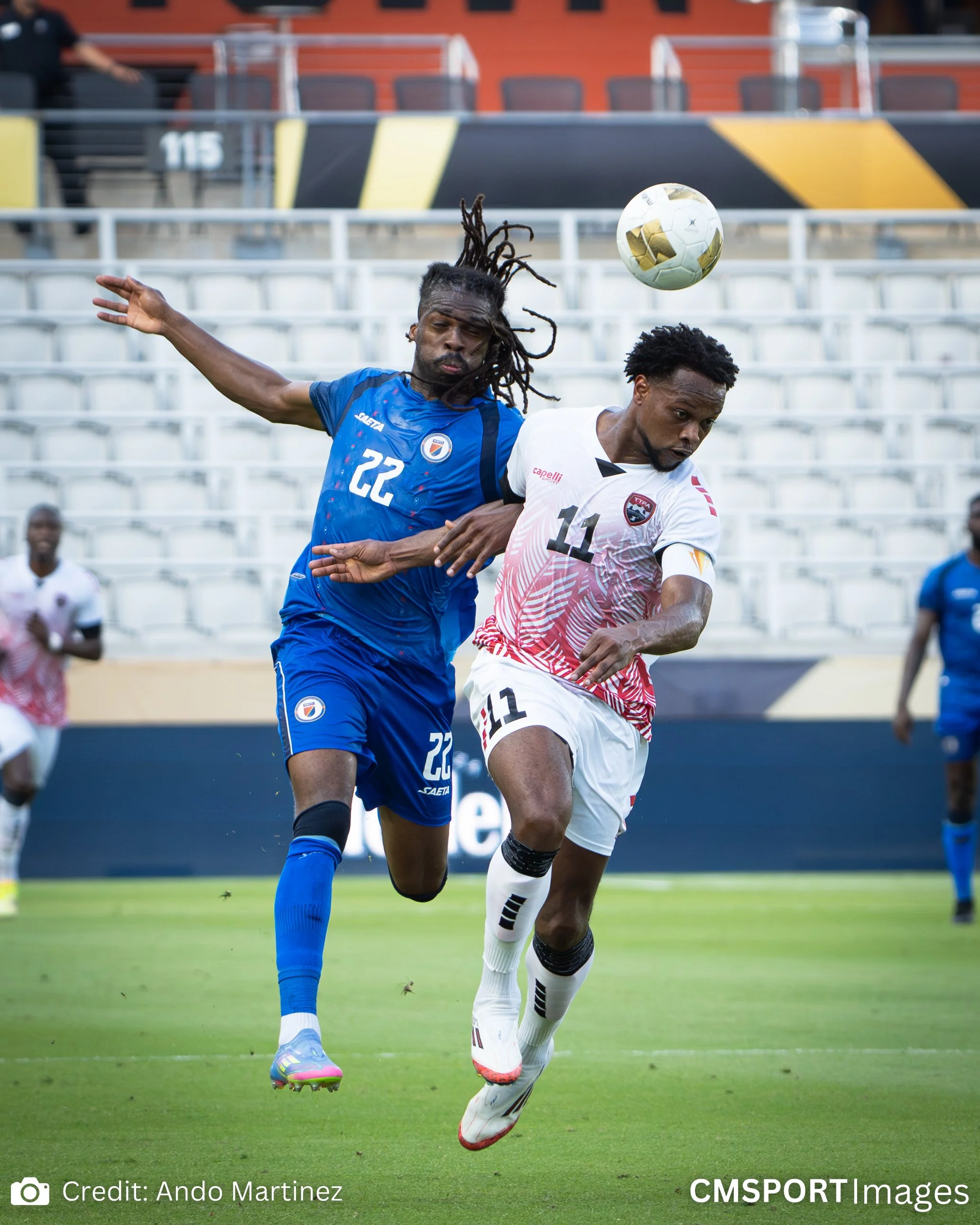 Two soccer players jump for the ball during a match. One player is wearing a blue jersey, and the other is wearing a white and red jersey. The background shows an empty stadium with white seats.