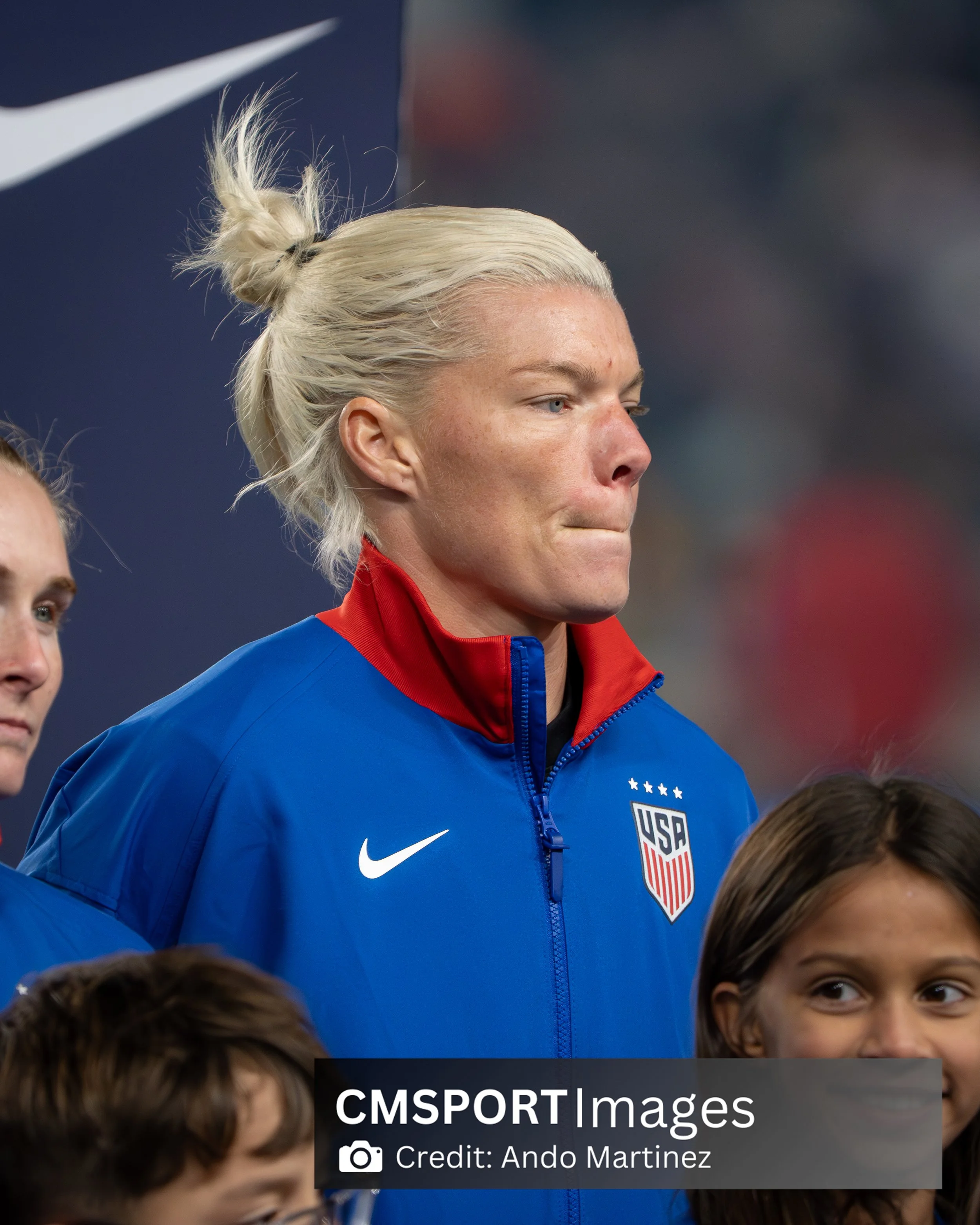 U.S. women's national soccer team player with platinum blonde hair tied in a bun, wearing a blue and red USA jacket, standing among children on a soccer field.