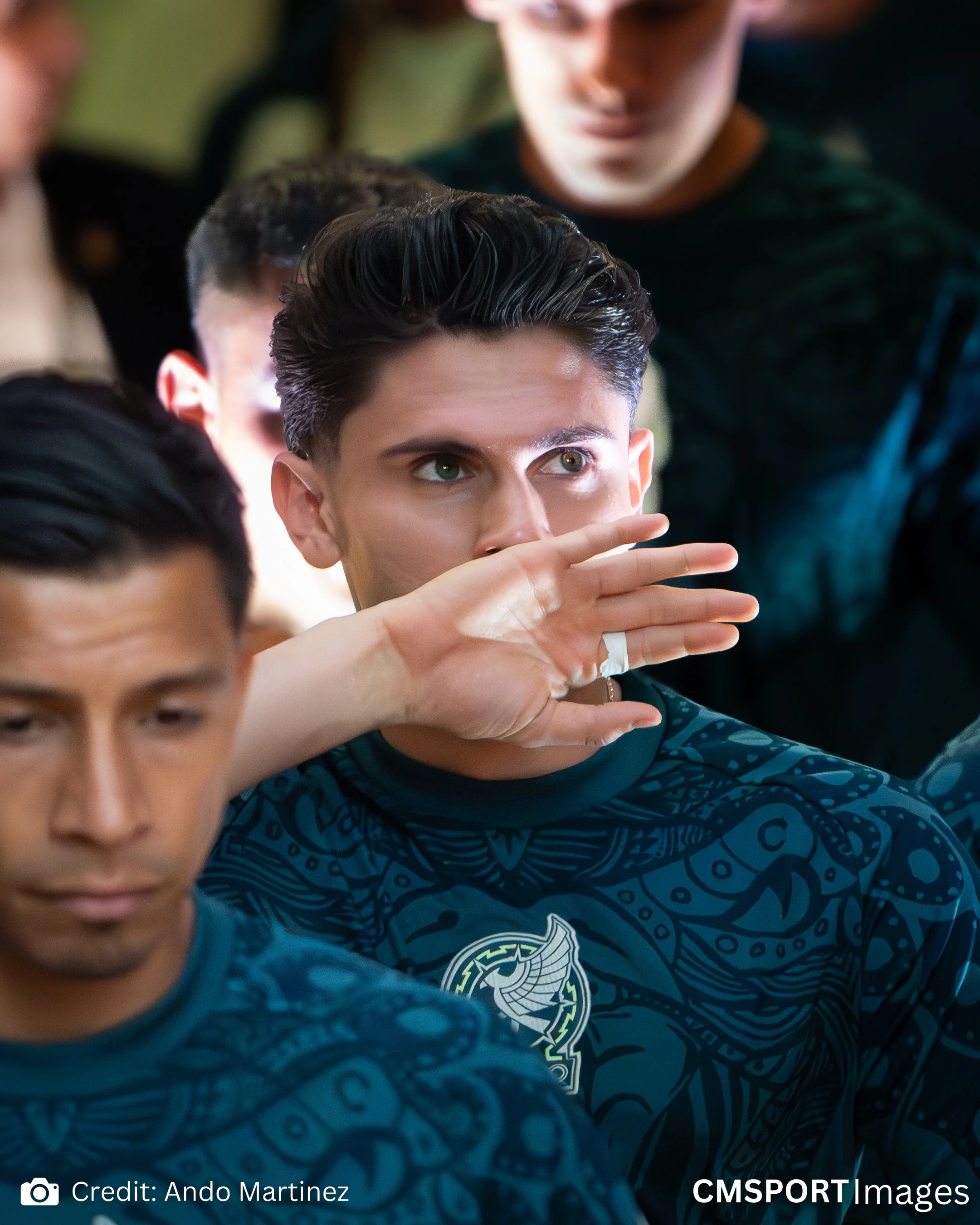 A group of young men, possibly athletes, dressed in dark sports jerseys with intricate designs, sitting closely together. One man in the center is raising his hand to his face, possibly to brush his nose or scratch it, and is wearing a ring on his fi