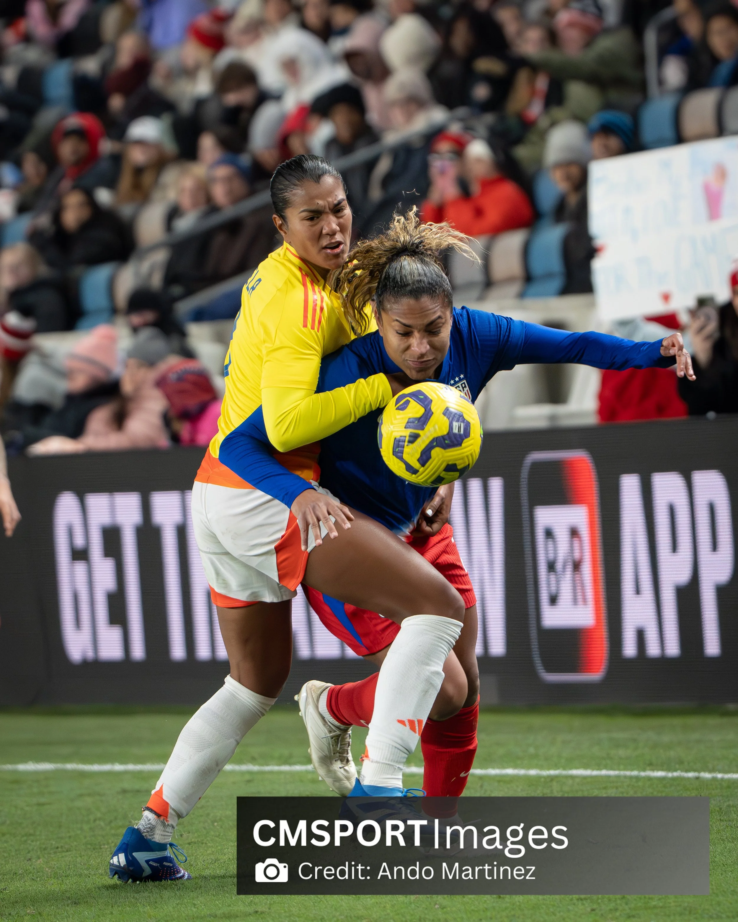 Two female soccer players competing for the ball during a match, one in a yellow jersey and the other in a blue jersey, on a soccer field with spectators in the background.