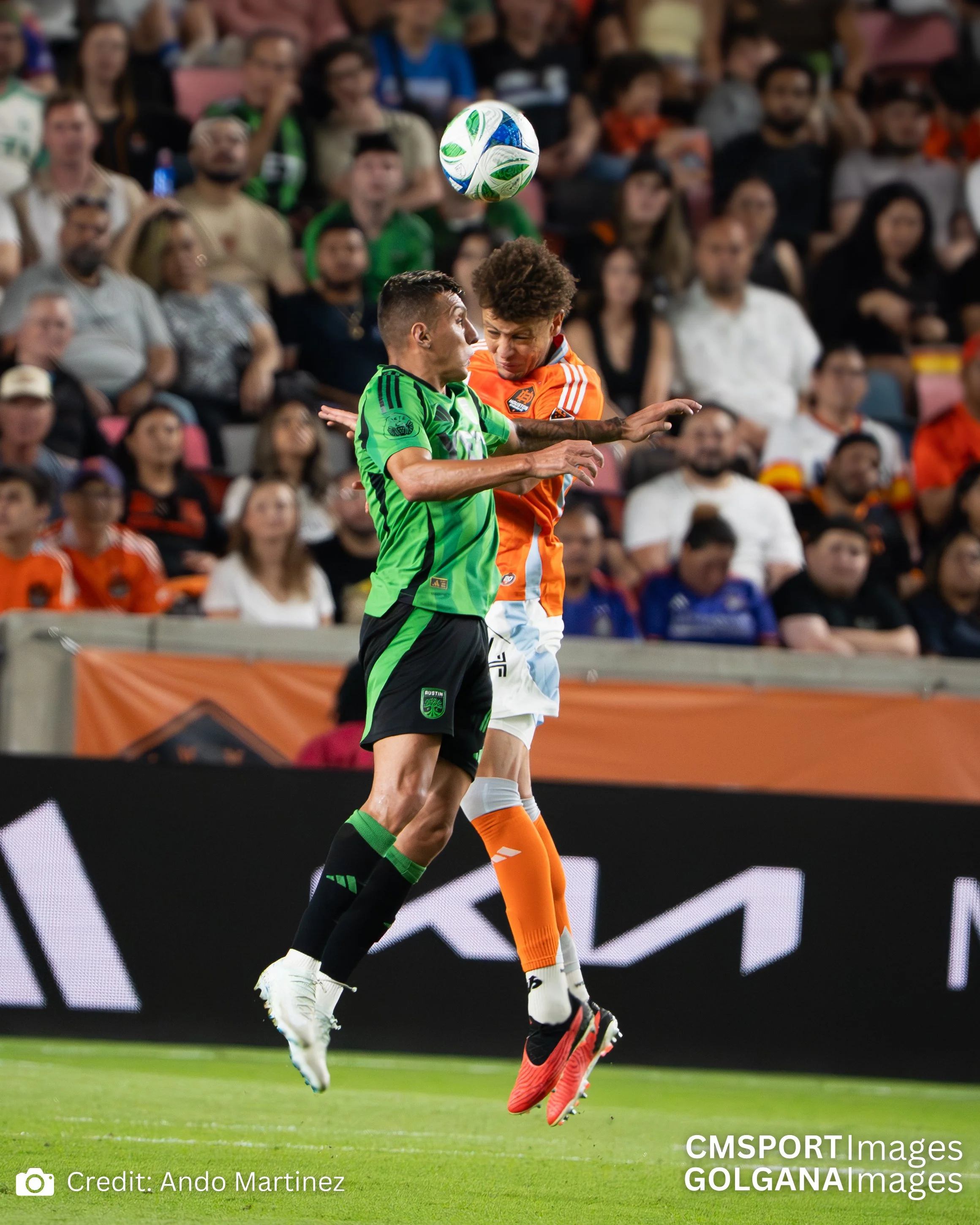Two soccer players, one in a green and black uniform and the other in an orange and white uniform, jump to head the soccer ball during a match, with a large crowd of spectators in the background.