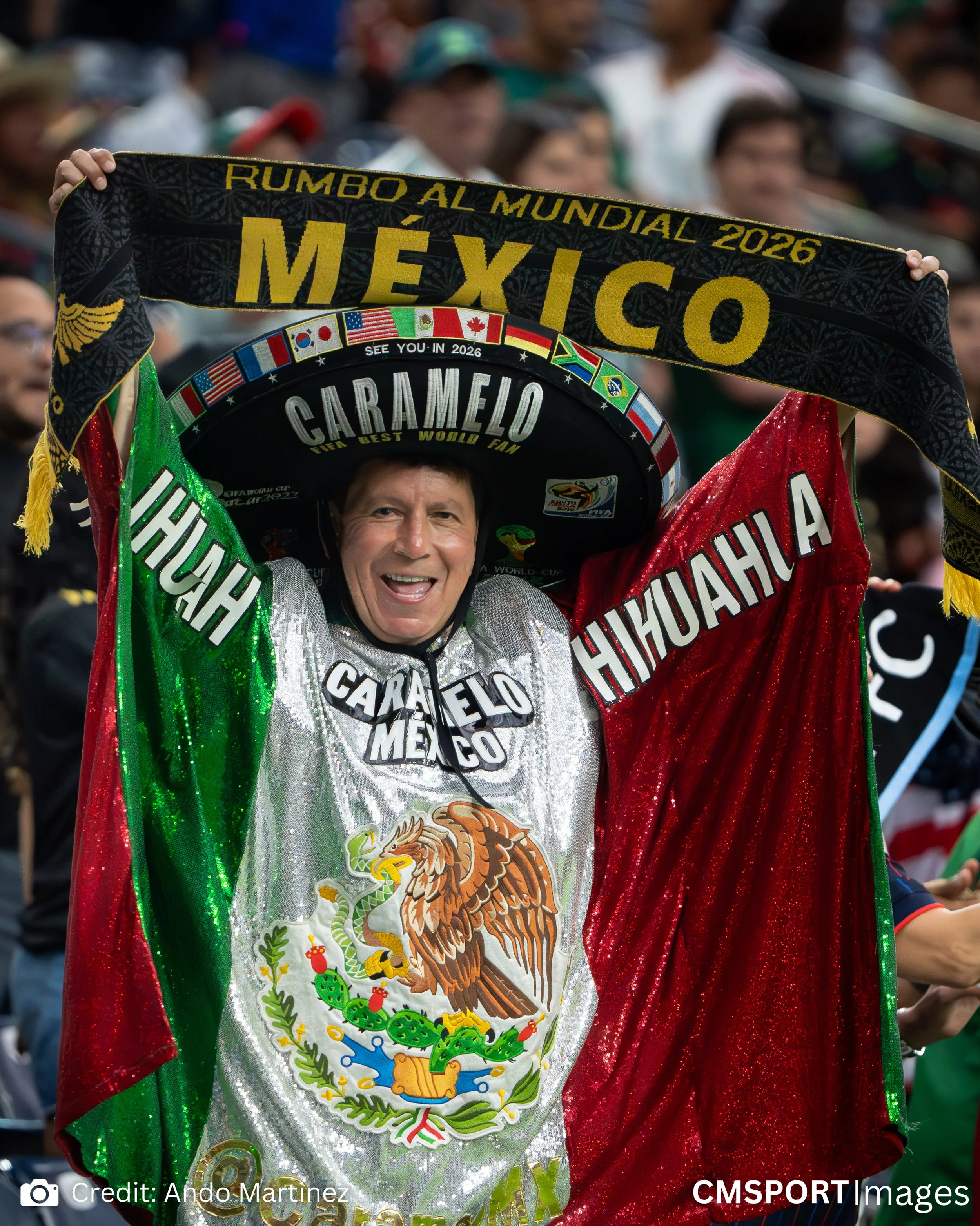 A Mexican soccer fan dressed in Mexican colors, holding a sombrero with flags, celebrating at a stadium, smiling, and draped in the Mexican flag with a large eagle emblem, during a sporting event.