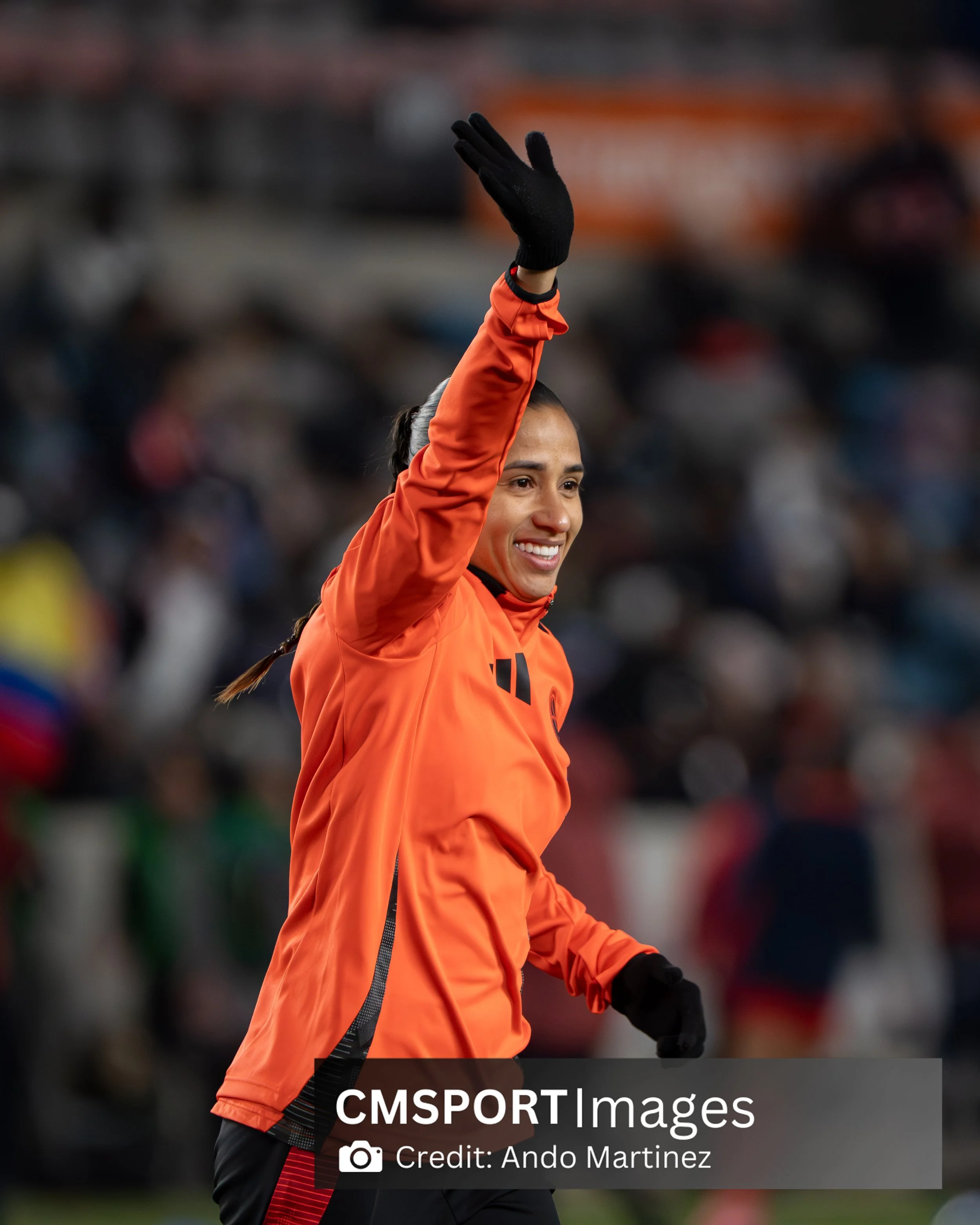 Female soccer player waving at the crowd, dressed in orange sportswear and black gloves, smiling at a stadium.