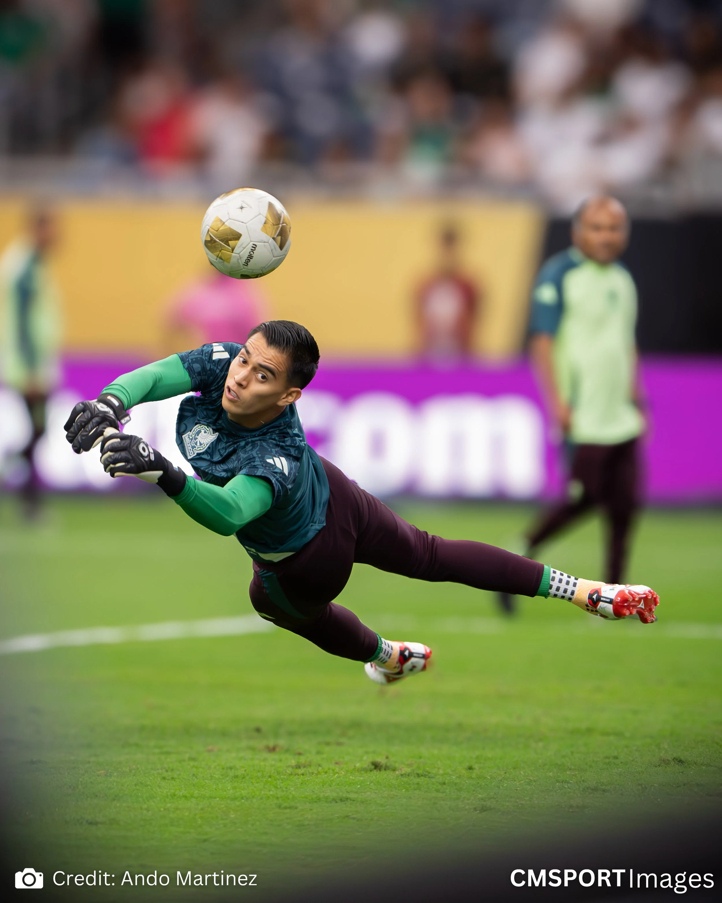 A soccer goalkeeper in a blue shirt and maroon pants dives to catch a soccer ball during a game in a stadium with spectators in the background.