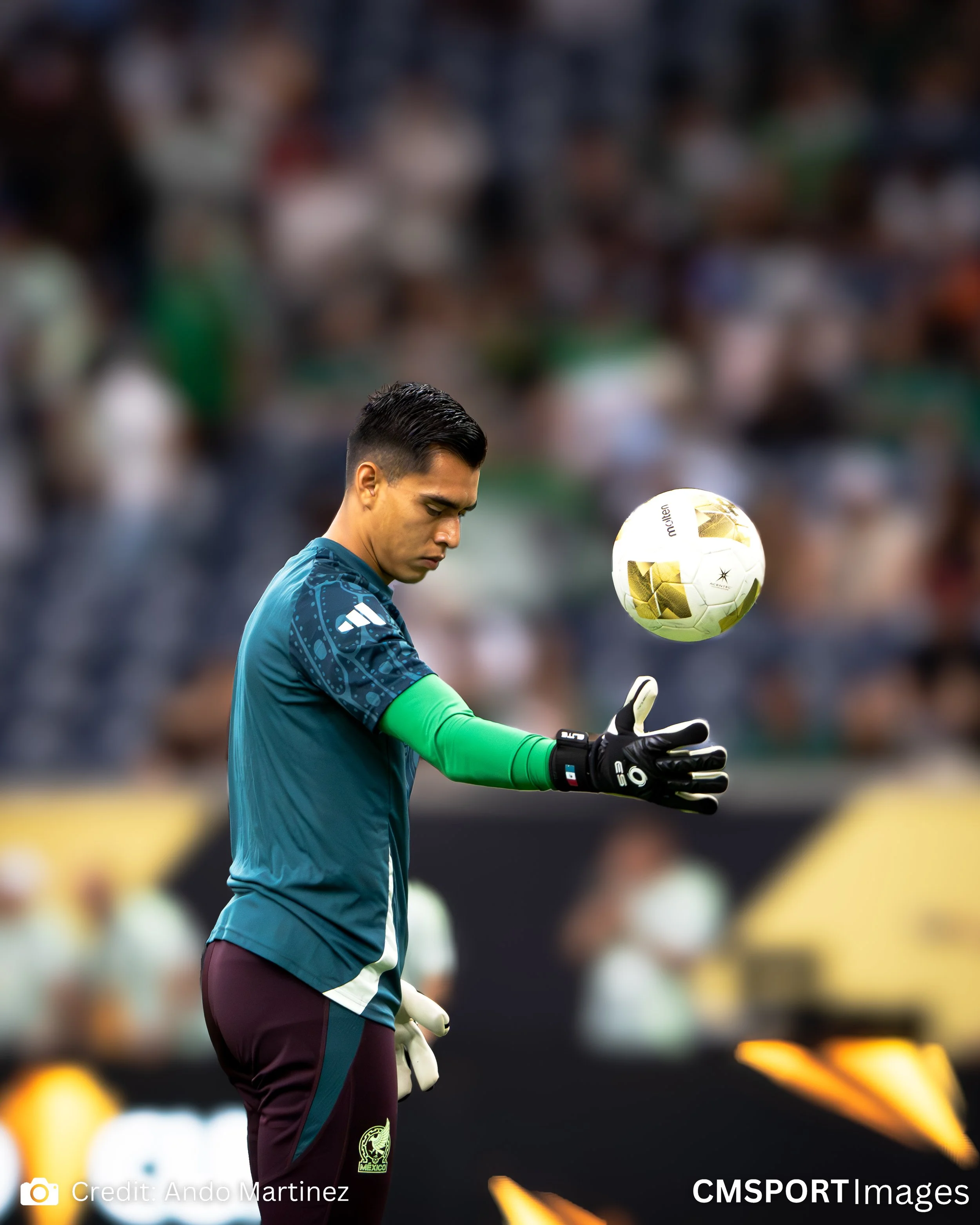 A soccer goalkeeper in teal uniform and black gloves is catching or holding a white and gold soccer ball during a game.