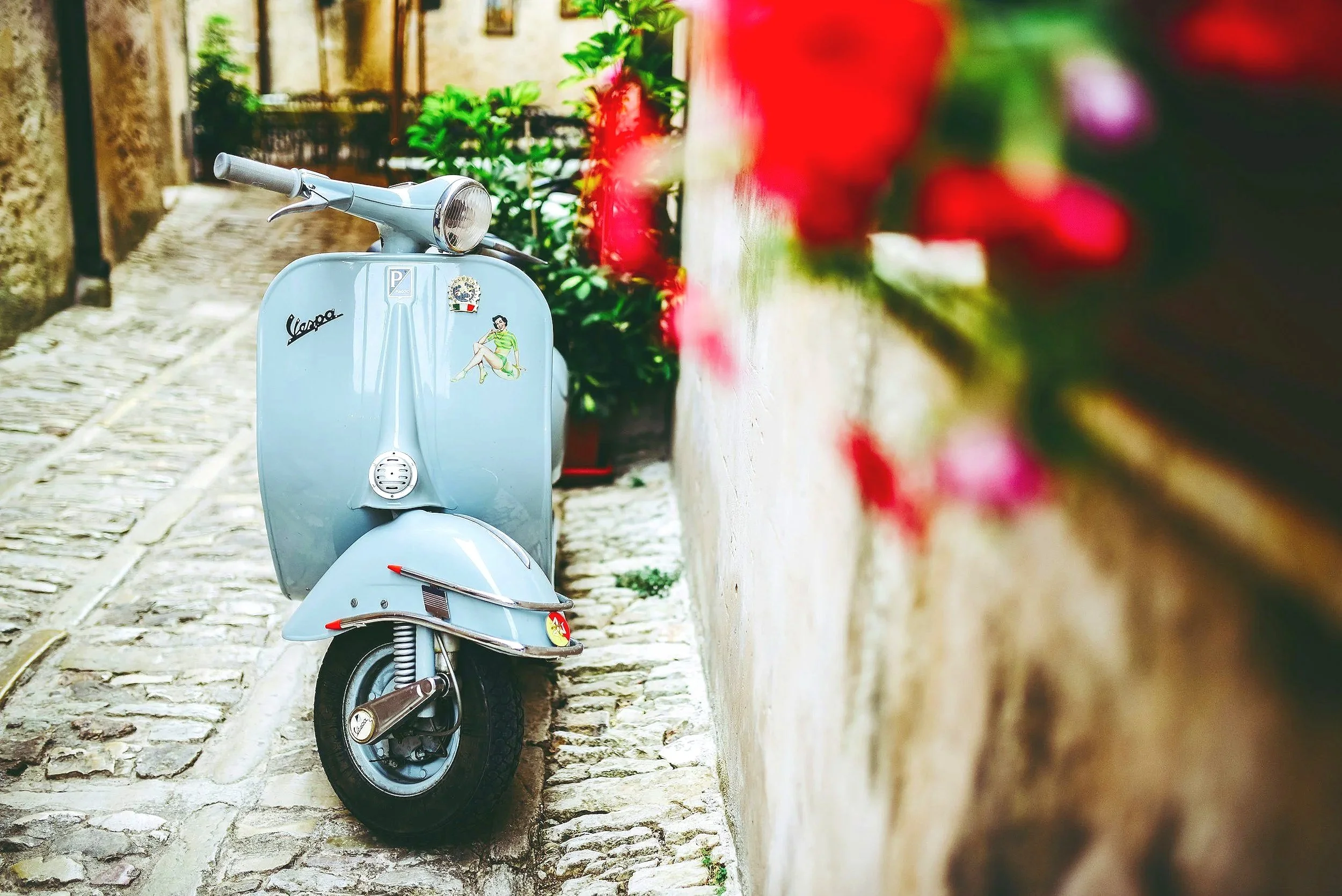 Vintage Vespa scooter parked on a narrow cobblestone alley with red flowers on the side.