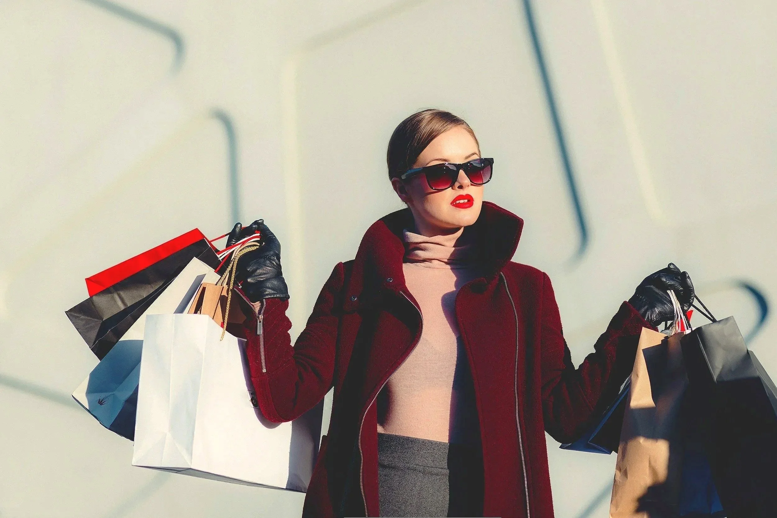 Fashionable woman with shopping bags, wearing a red coat and sunglasses outdoors.