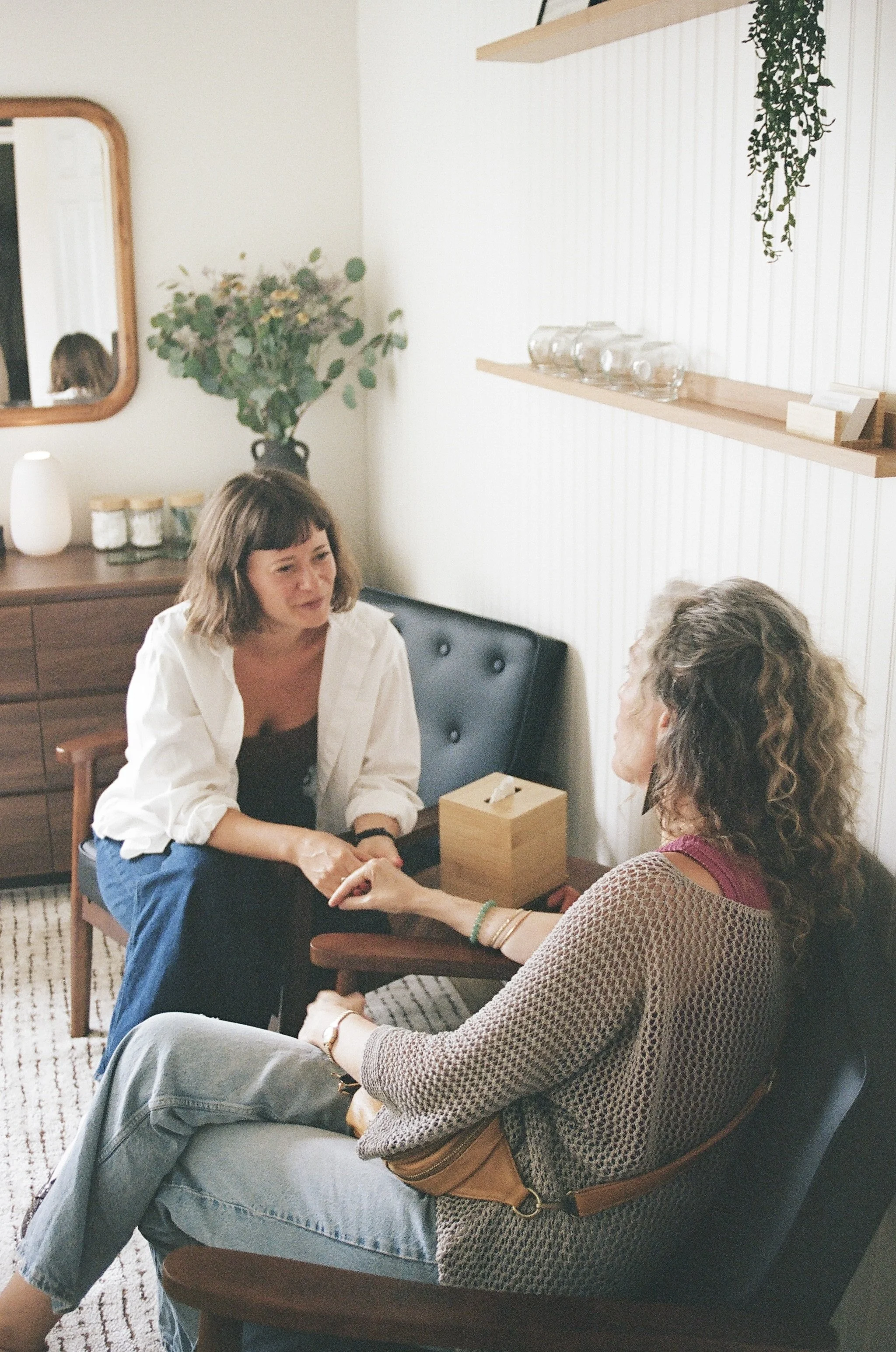 Two women are sitting on chairs in a cozy, well-decorated room. They are holding hands and engaging in a heartfelt conversation. One woman, with shoulder-length brown hair, is wearing a white shirt and blue jeans. The other woman, with curly hair, is dressed in a patterned sweater and gray pants, with a brown purse resting on her lap. On the side, there's a wooden sideboard with glass jars, a white diffuser, and a mirror on the wall. A large vase with greenery is placed on a shelf, adding to the room's warm ambiance.