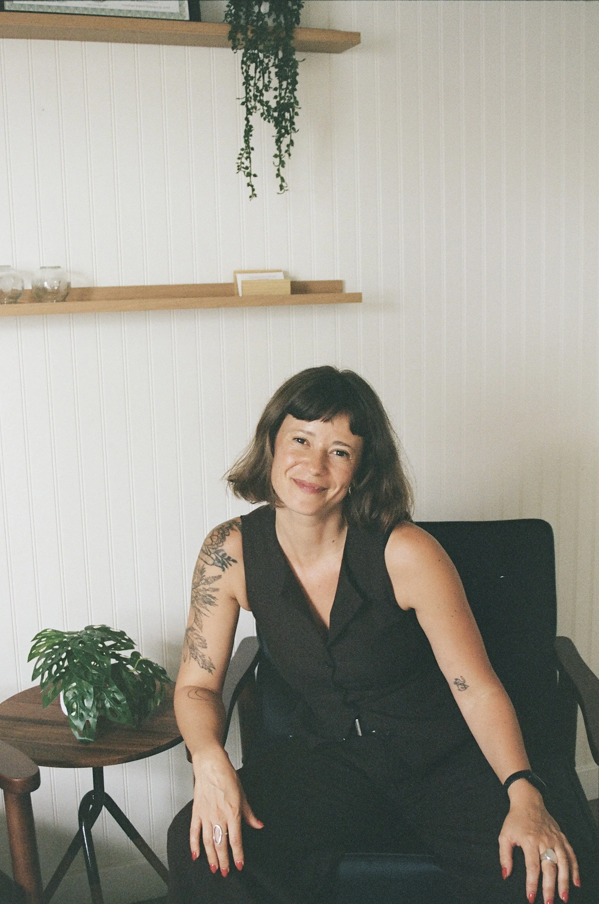A woman with brown wavy hair, tattoos on her arms, wearing a black sleeveless top, seated at a desk with a plant on her left, smiling at the camera, against a white paneled wall with wooden shelves and a hanging green plant.