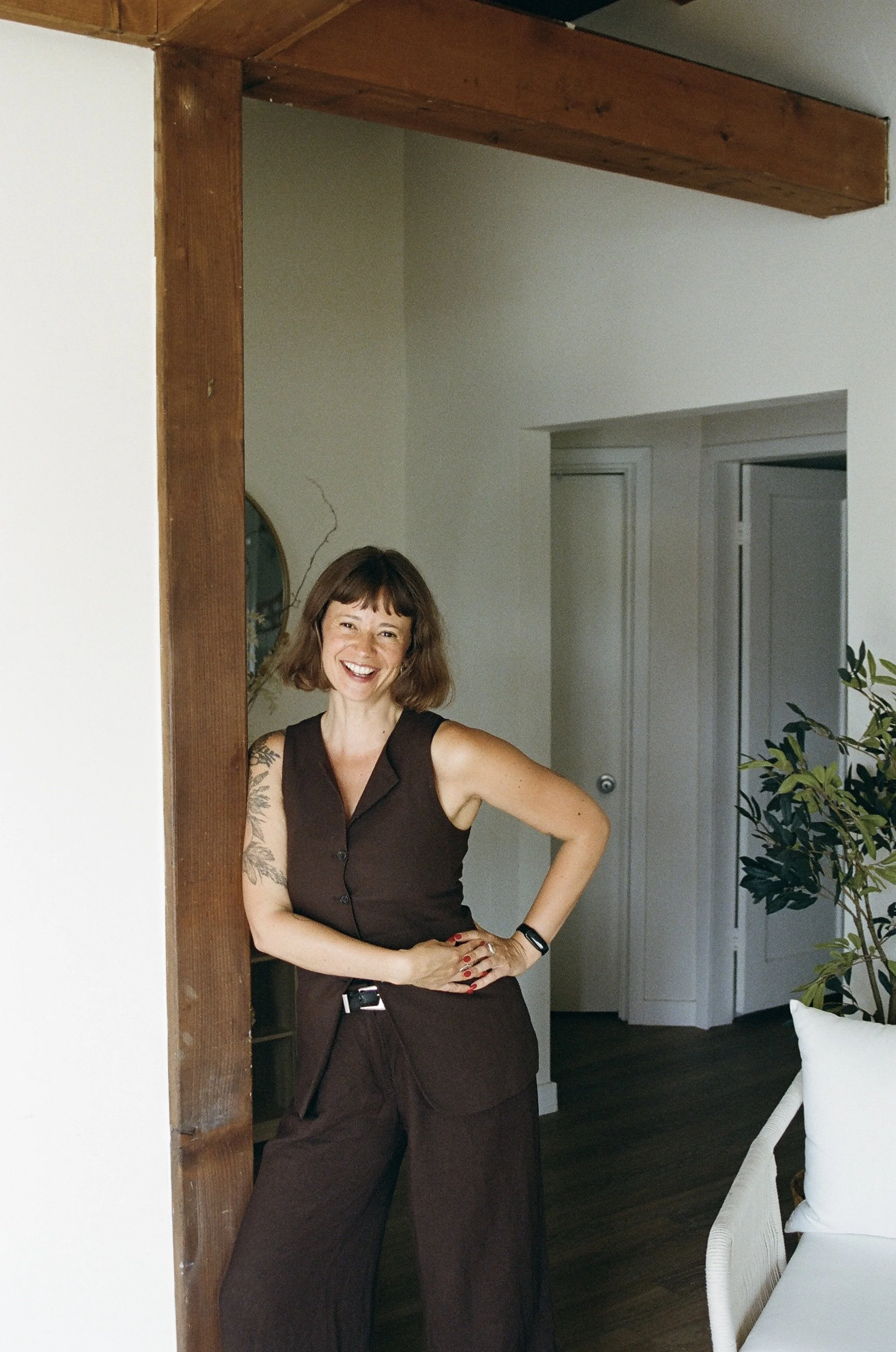 A woman with short brown hair and tattoos on her left arm, wearing a sleeveless brown top and wide-leg brown pants, standing indoors near a white wall and a wooden beam, smiling with her hands on her hips, next to a white chair with a woven pattern, in a room with white walls, doors, and a plant.