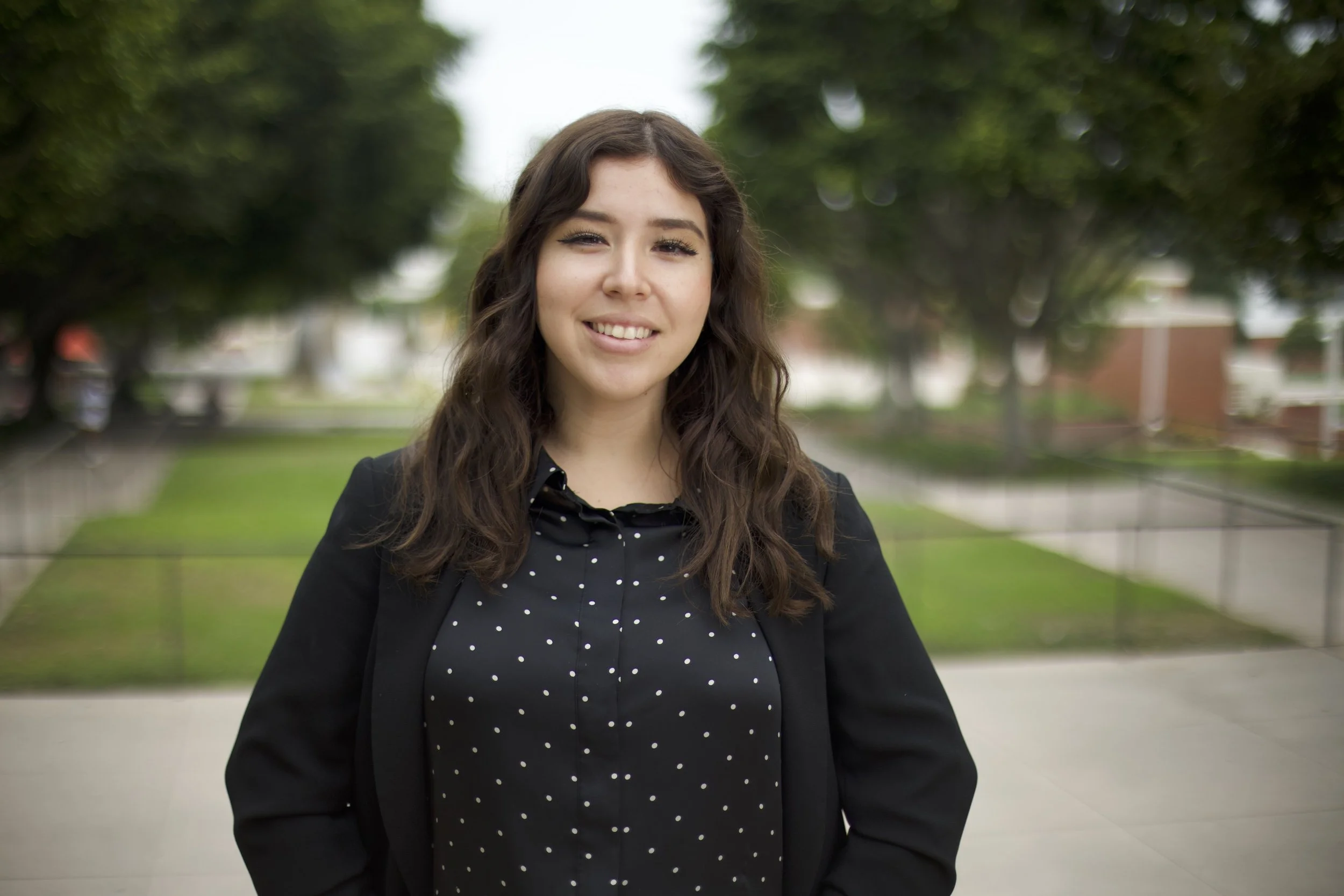 A young woman with wavy brown hair, wearing a black blazer and a black polka dot blouse, standing outdoors on a cloudy day with a smile.
