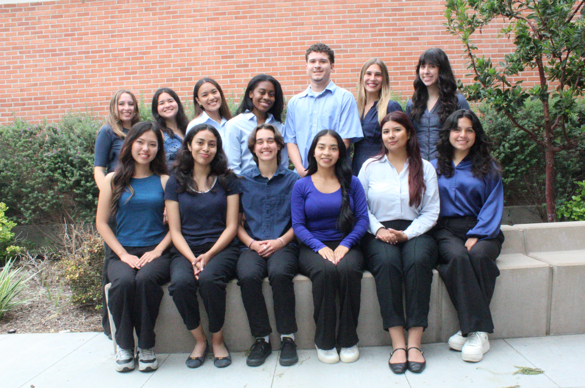 Group of fourteen young adults assembled outdoors, sitting and standing in front of a brick wall and green bushes, posing for a photo.