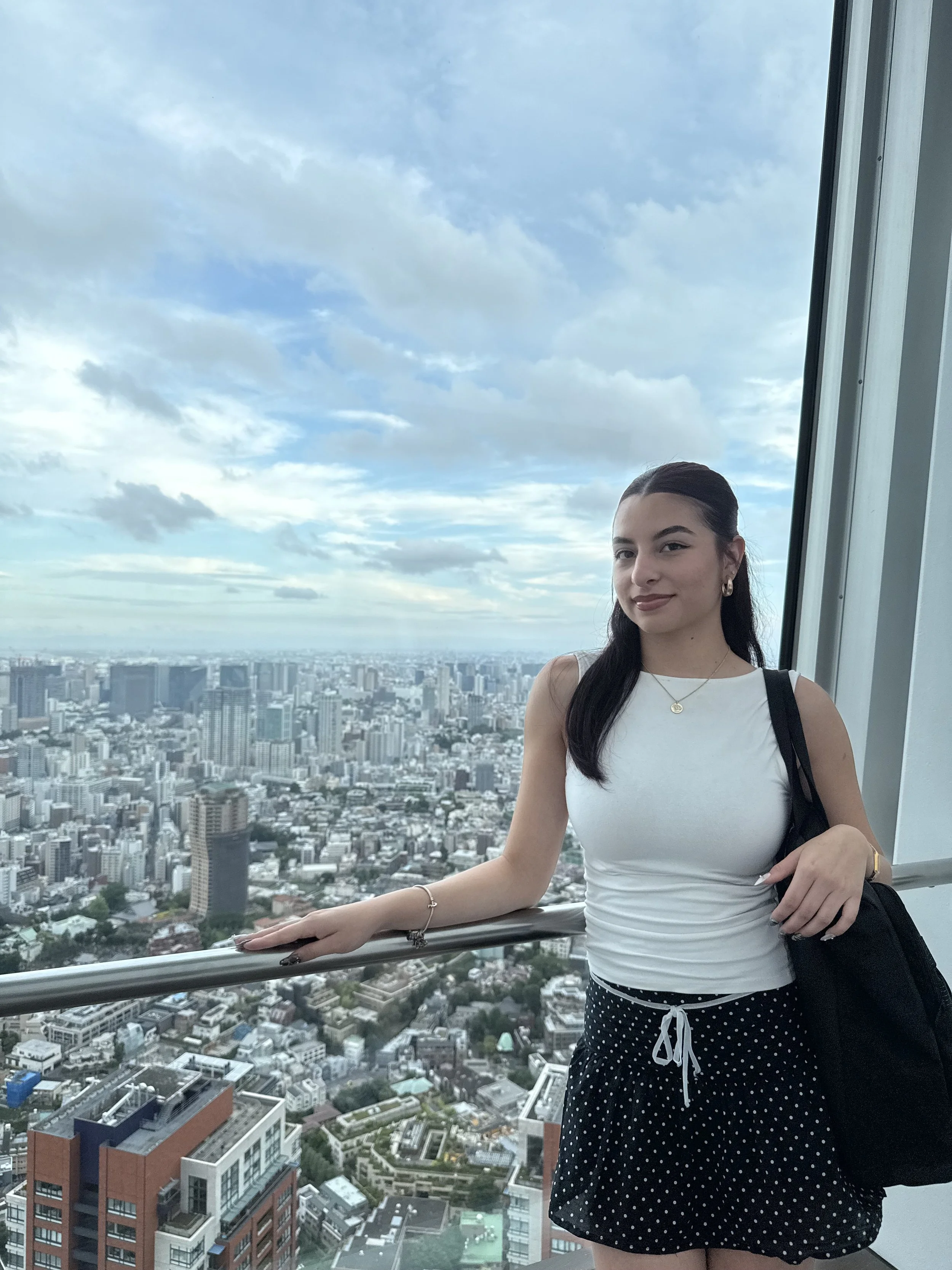 A young woman with dark hair, wearing a white tank top and polka dot shorts, stands on a high observation deck overlooking a city skyline with tall buildings and cloudy sky.