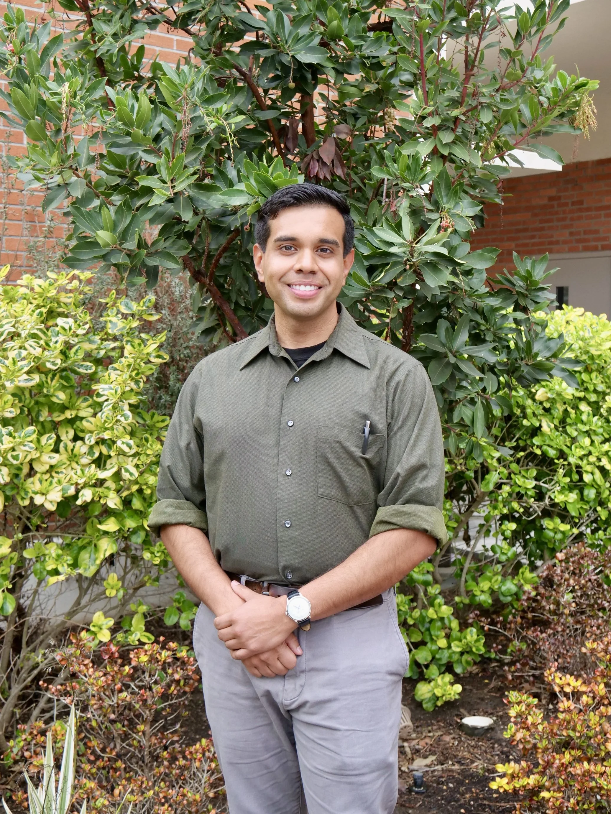 A smiling man in a green shirt and gray pants standing outdoors in front of lush green bushes and trees.