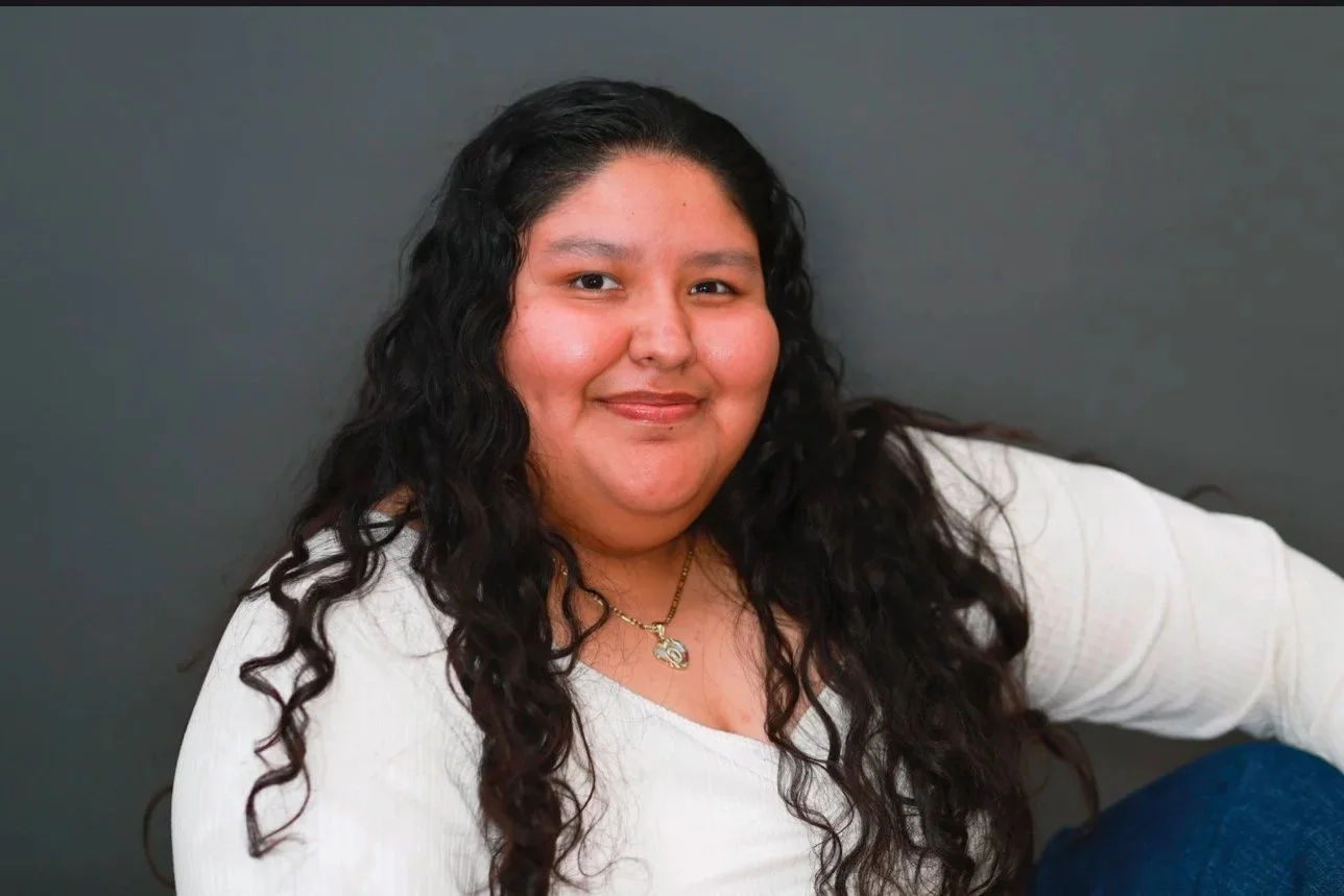 A woman with long, dark, curly hair sitting against a plain gray wall, smiling at the camera, wearing a white top and a gold necklace.