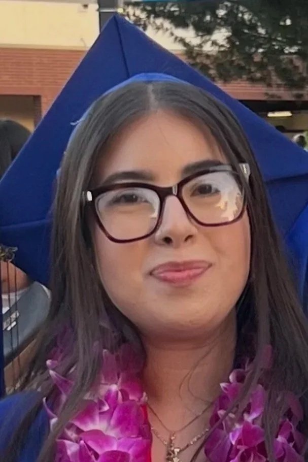 A young woman wearing glasses and a blue graduation cap, standing outdoors during daytime, with a purple flower lei around her neck.