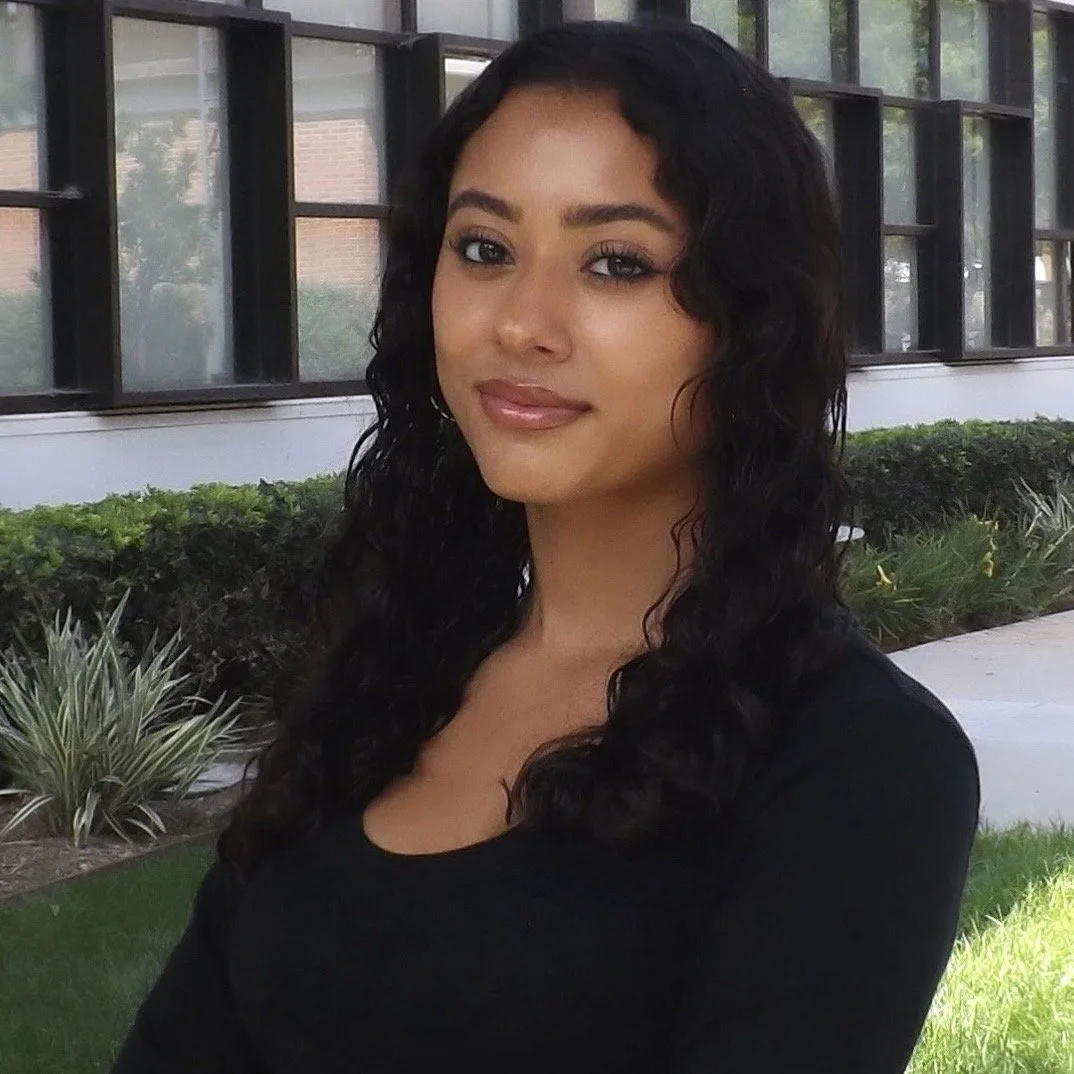 A young woman with dark, curly hair and tan skin stands outdoors in front of a building with large windows and greenery. She is smiling softly and wearing a black top.