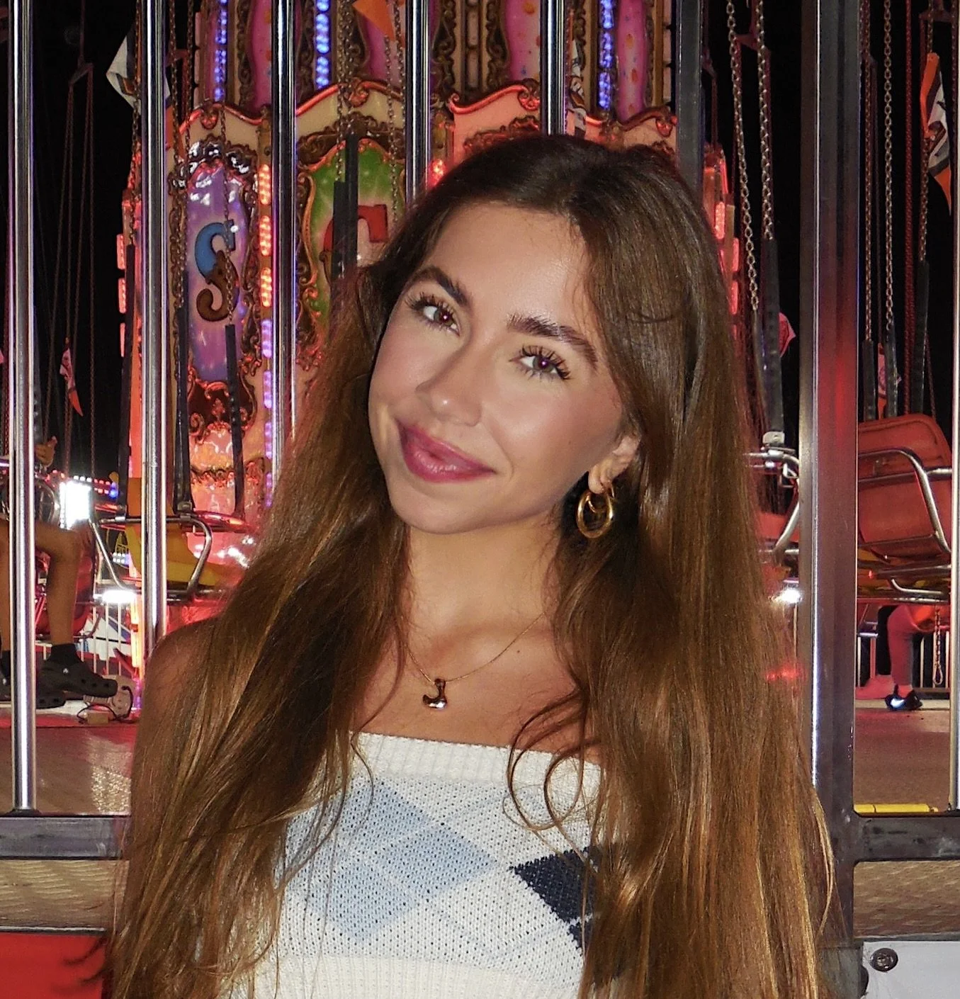 Young woman with long brown hair smiling at a carnival ride with colorful lights in the background.