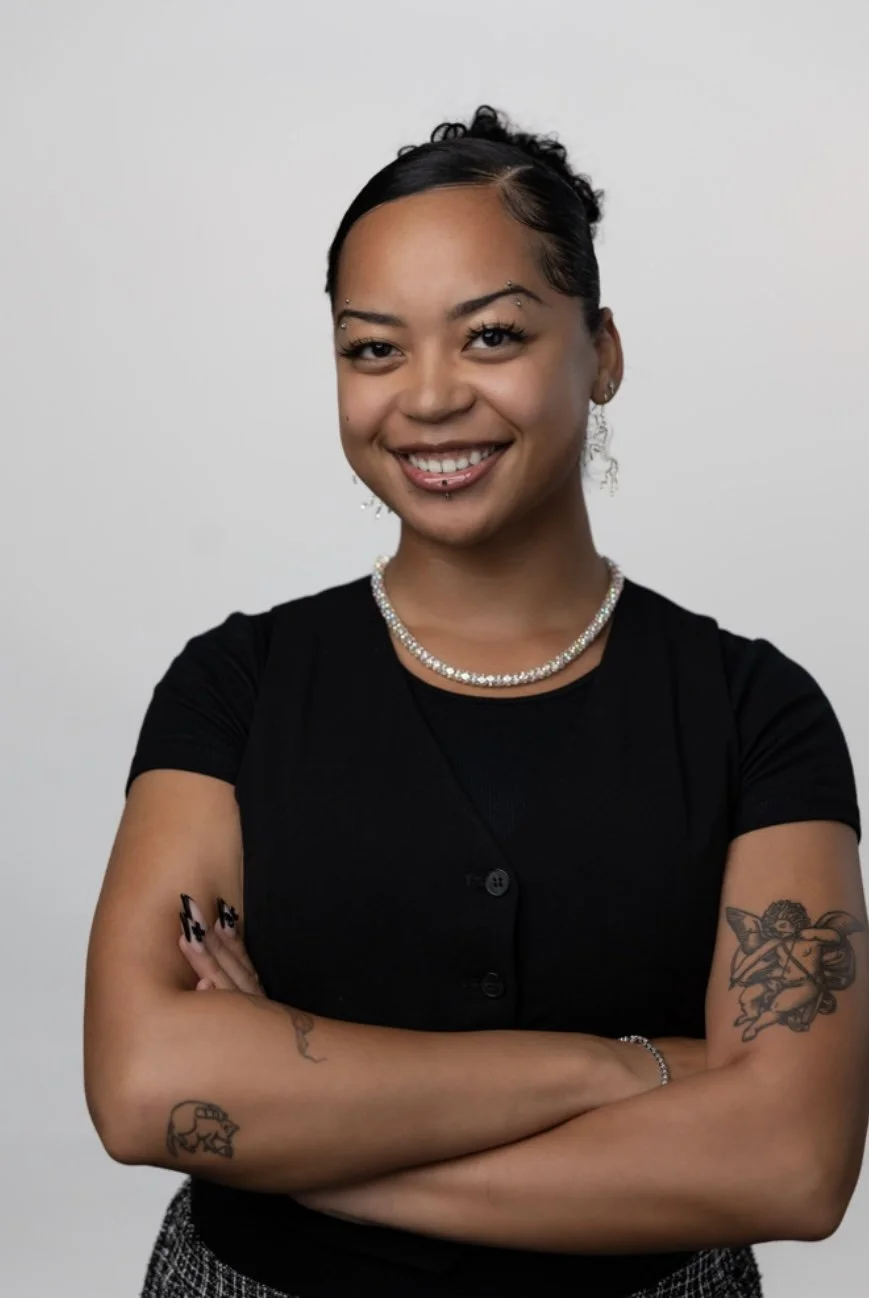 Portrait of a smiling young woman with tattoos, wearing earrings, a pearl necklace, and a black top, standing against a plain white background.