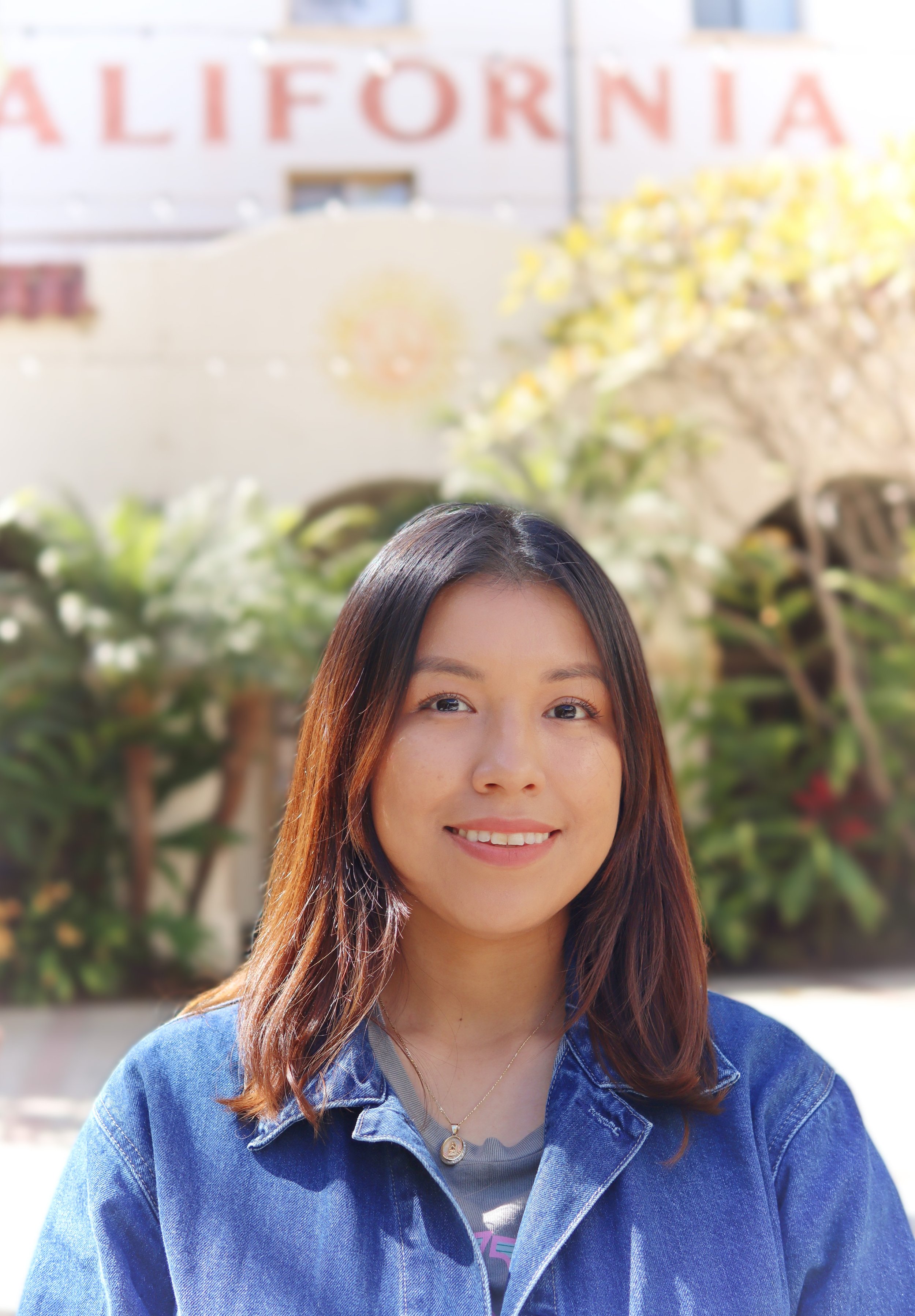 A young woman smiling outdoors with plants and a building with partially visible 'California' sign in the background.