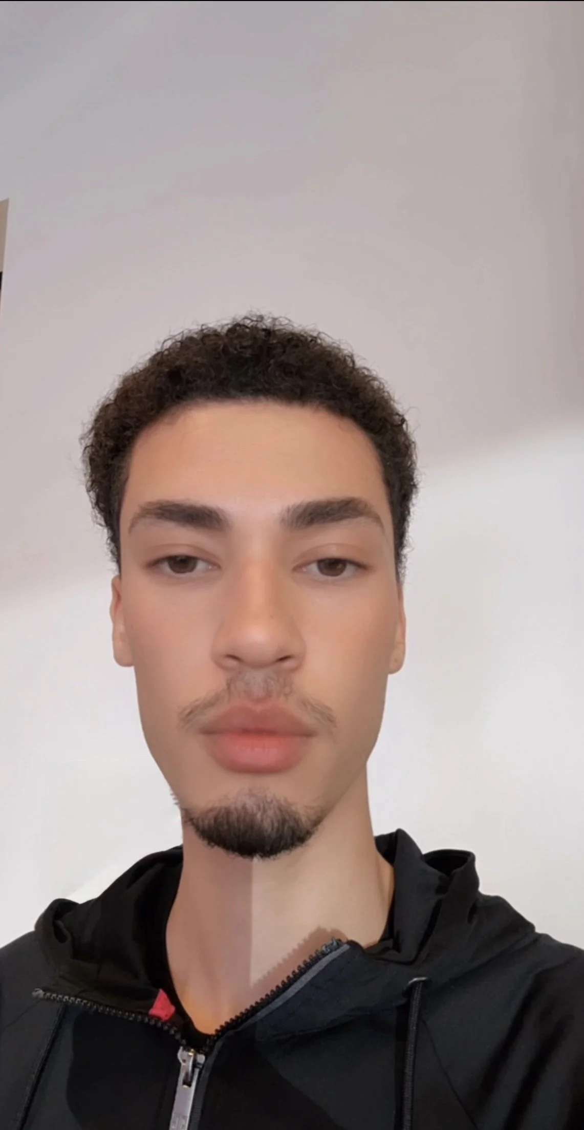 Close-up of a young man with short curly hair, light facial hair, and light skin, wearing a black zip-up hoodie, standing in front of a plain white wall.