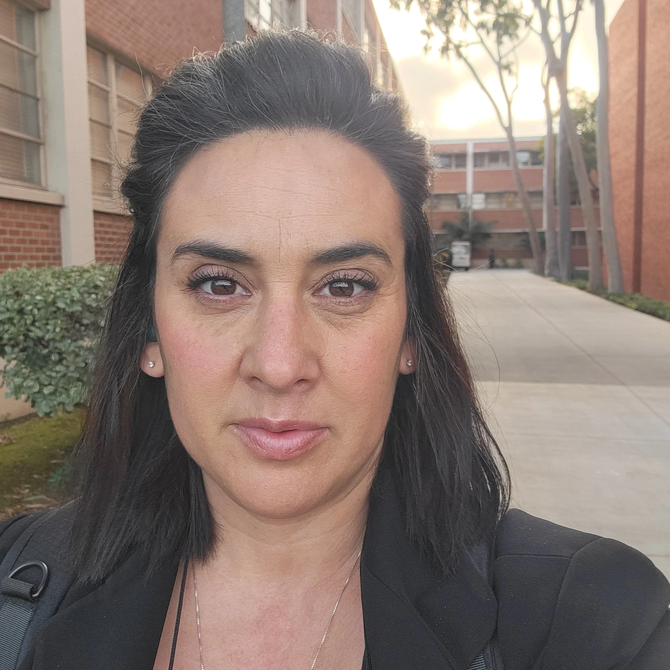 A woman with dark hair and light makeup posing outdoors near a university or office building, with trees and a walkway in the background.