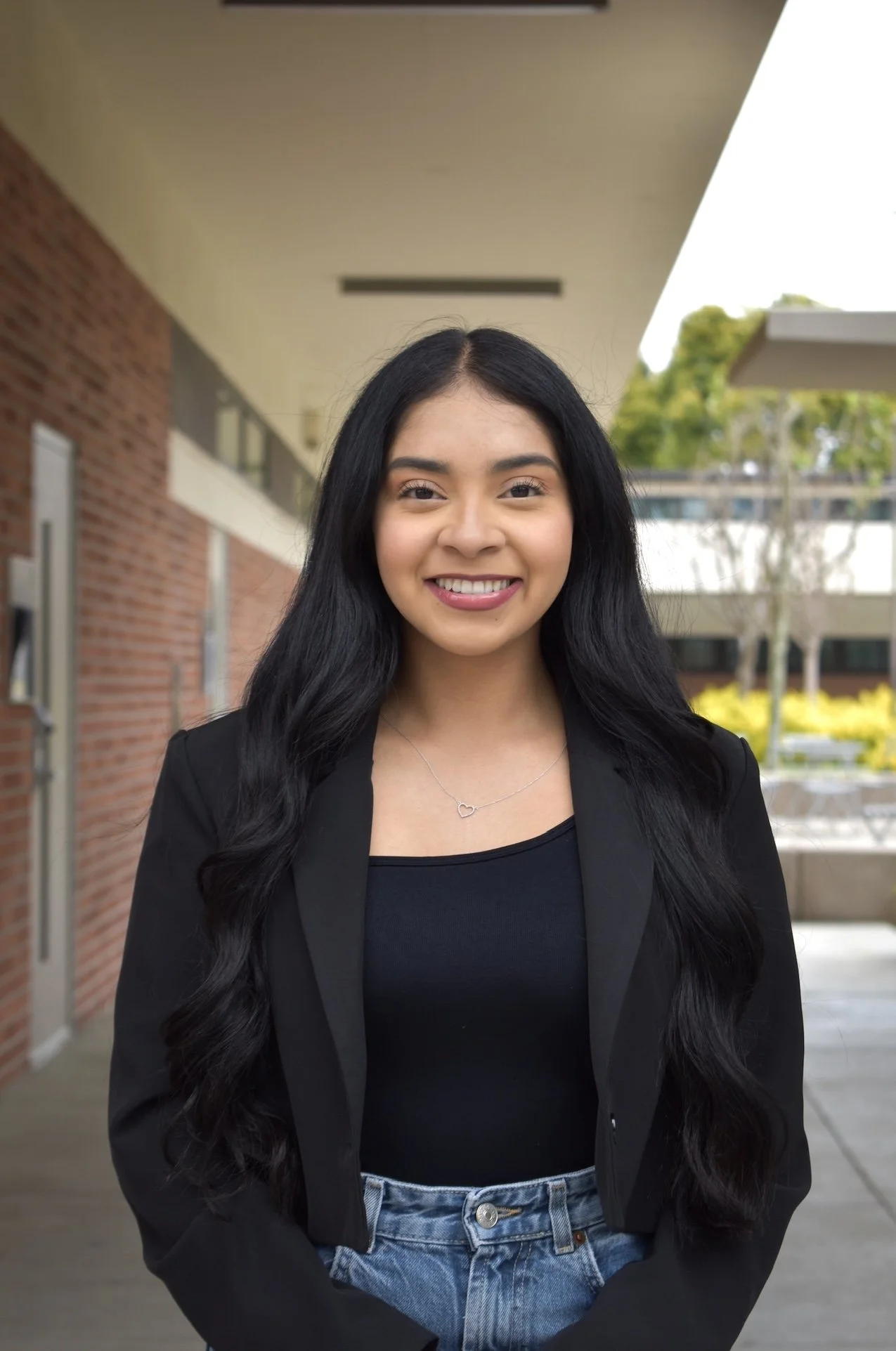 A young woman with long black hair, wearing a black blazer and a black top, smiling outdoors with a brick building and trees in the background.