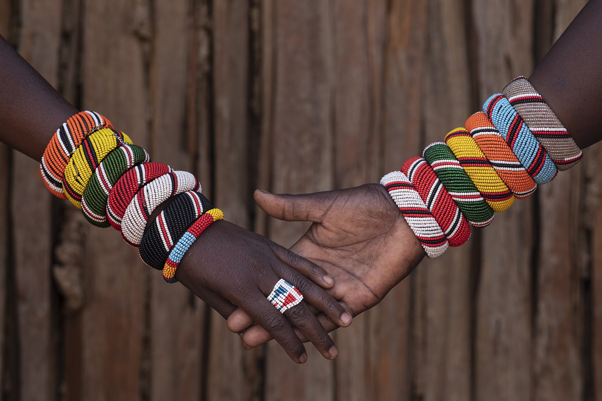 Two people wearing colorful beaded bracelets and a beaded ring, with hands clasped against a wooden background.
