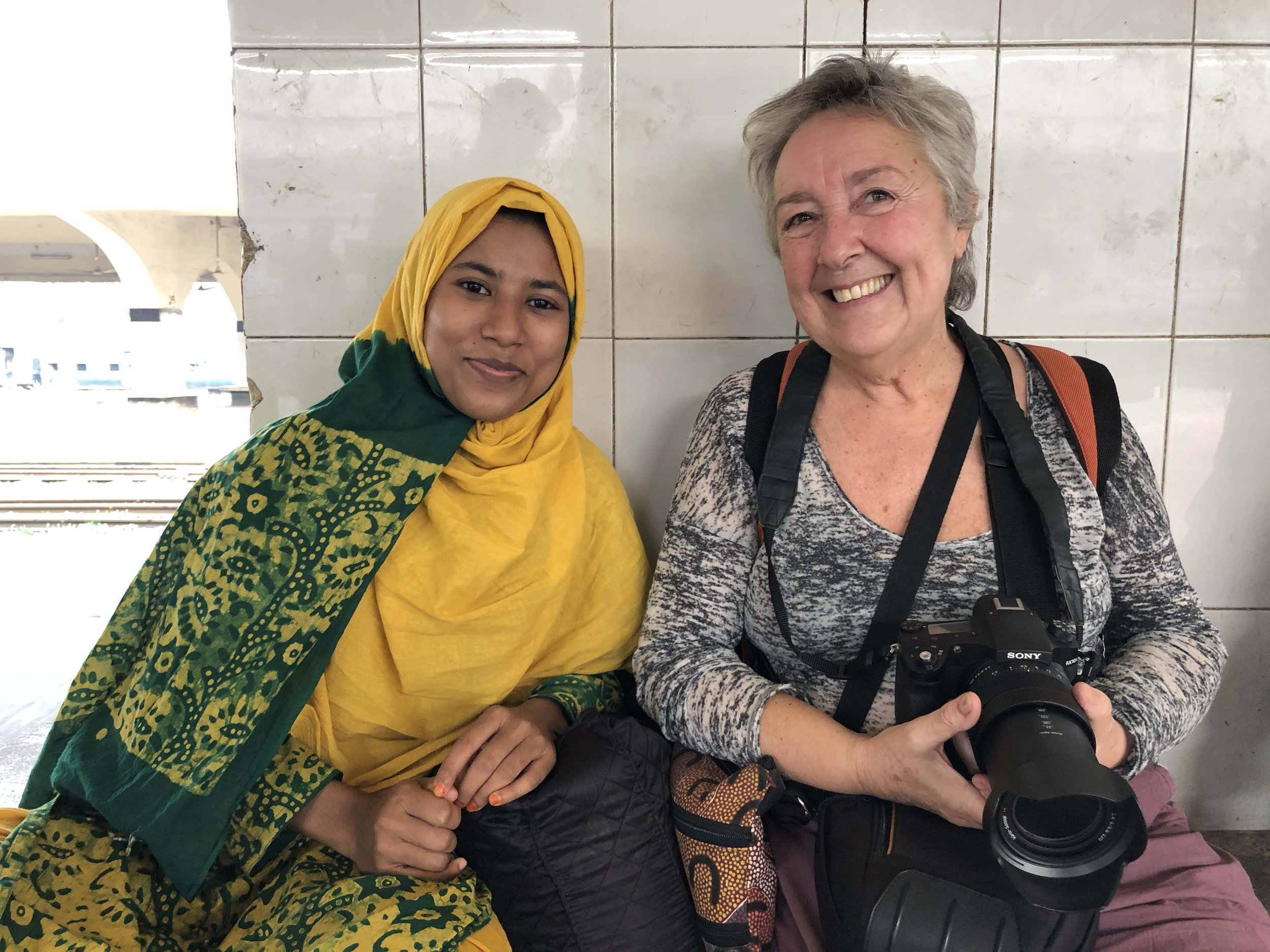 Two women sitting together, one wearing a yellow and green patterned headscarf and the other with short grey hair holding a Sony camera, smiling, against a tiled wall background.