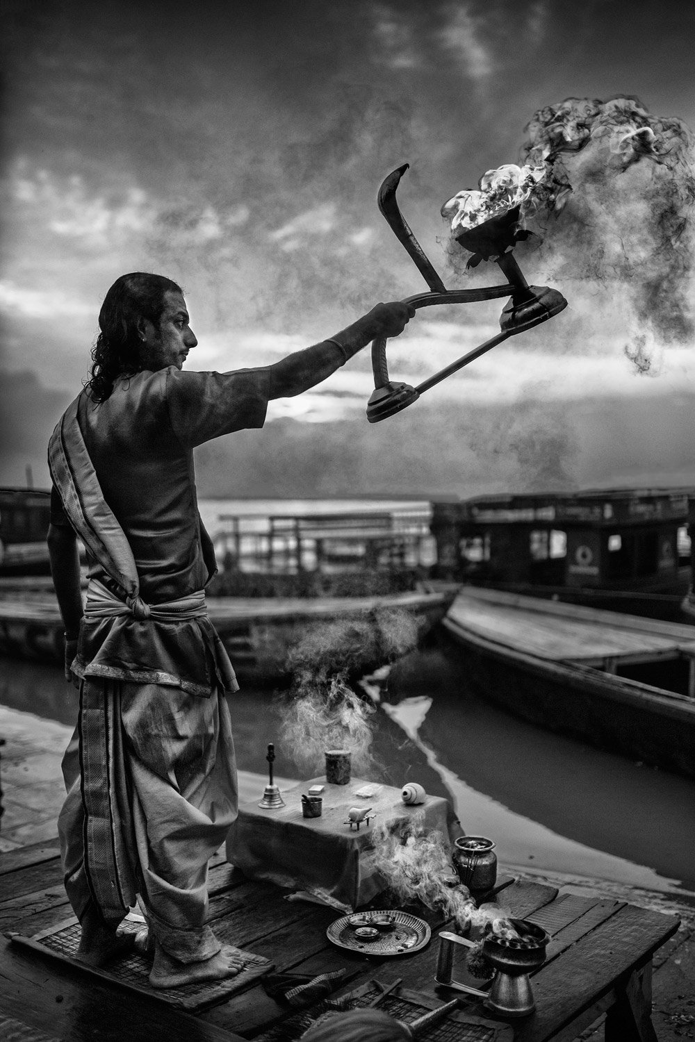 A person performing a traditional Hindu ritual by the river with a large incense holder, surrounded by ceremonial items on a wooden platform.