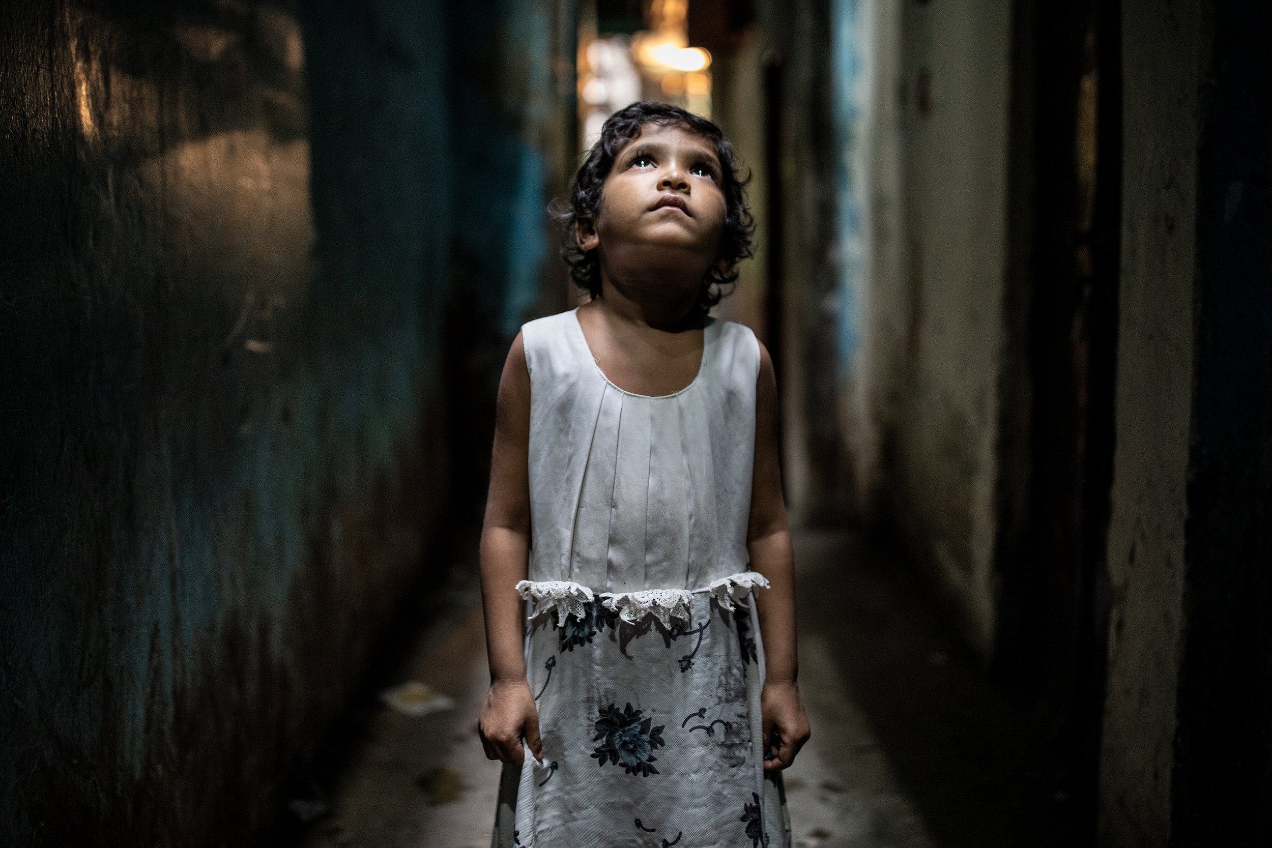 Child in a white dress standing in a narrow dimly lit alley, looking upward.