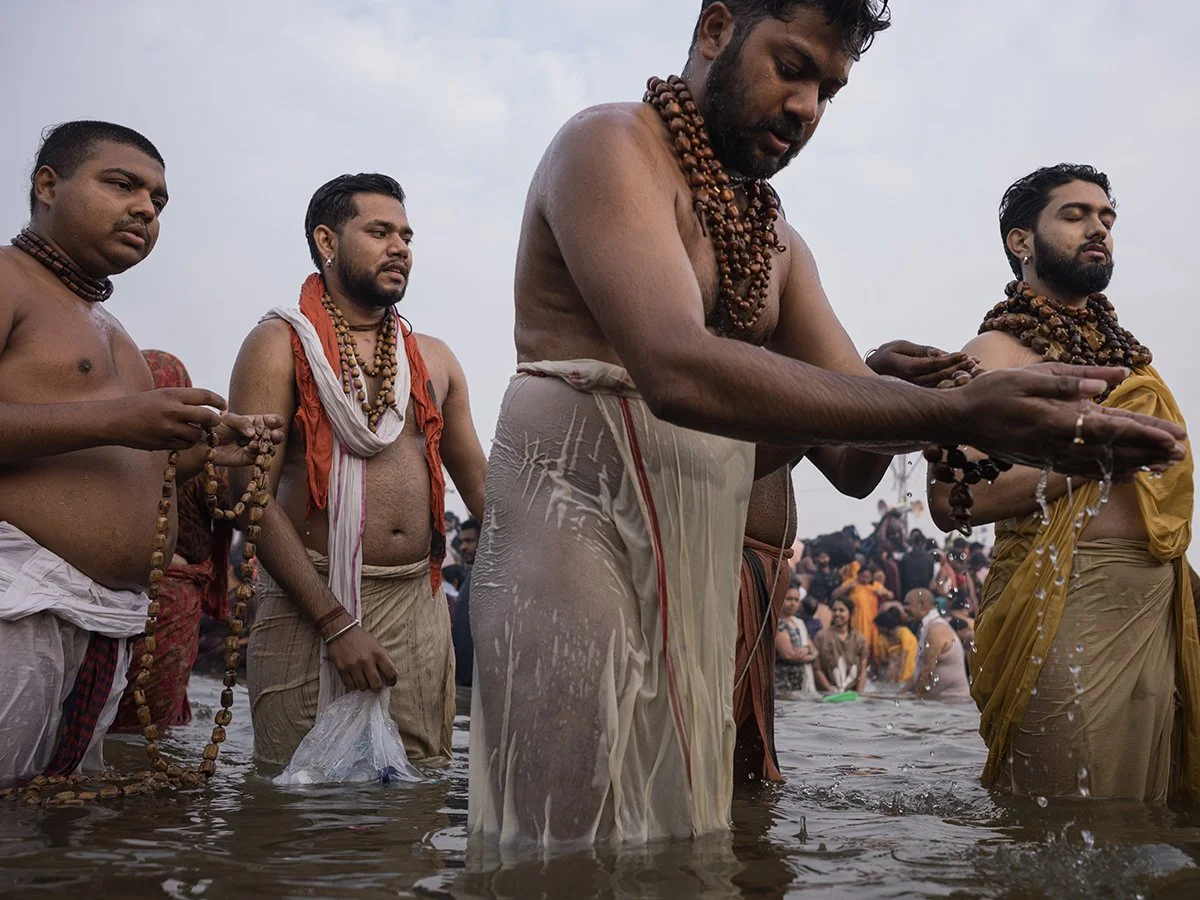 Men in traditional attire performing a ritual in water, wearing beaded necklaces, with a crowd in the background.